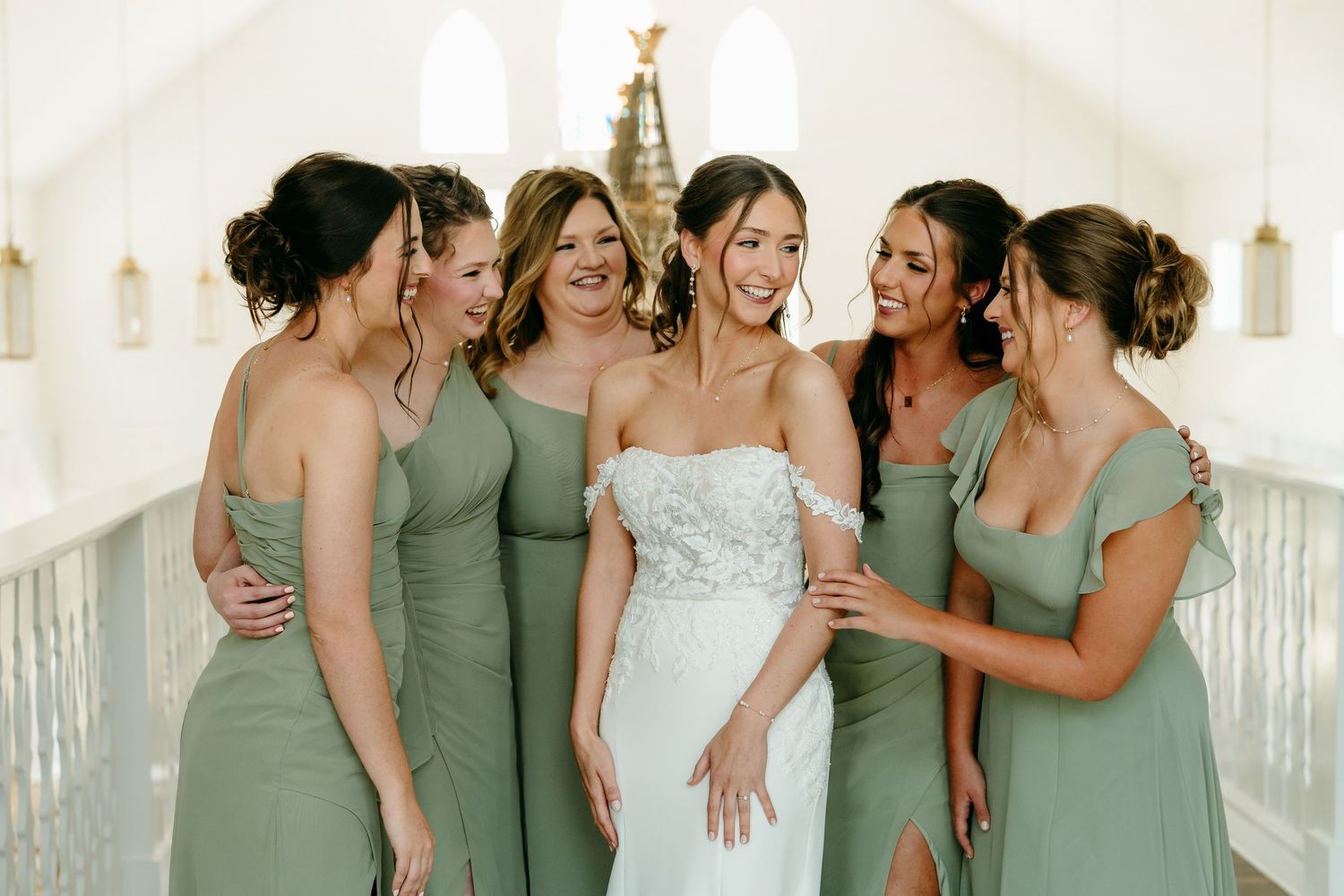 A wedding party in sage green bridesmaid dresses and a white wedding gown laugh together on a bright porch.