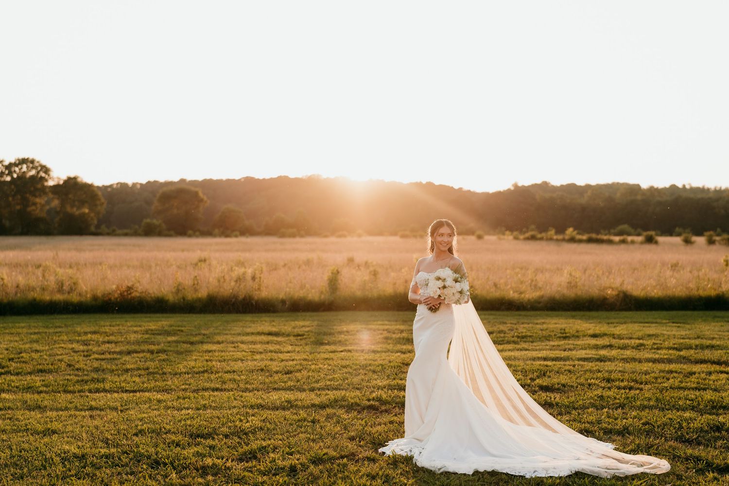 A bride in a flowing white gown stands in a sunlit field during golden hour as her veil catches the breeze Nashville TN