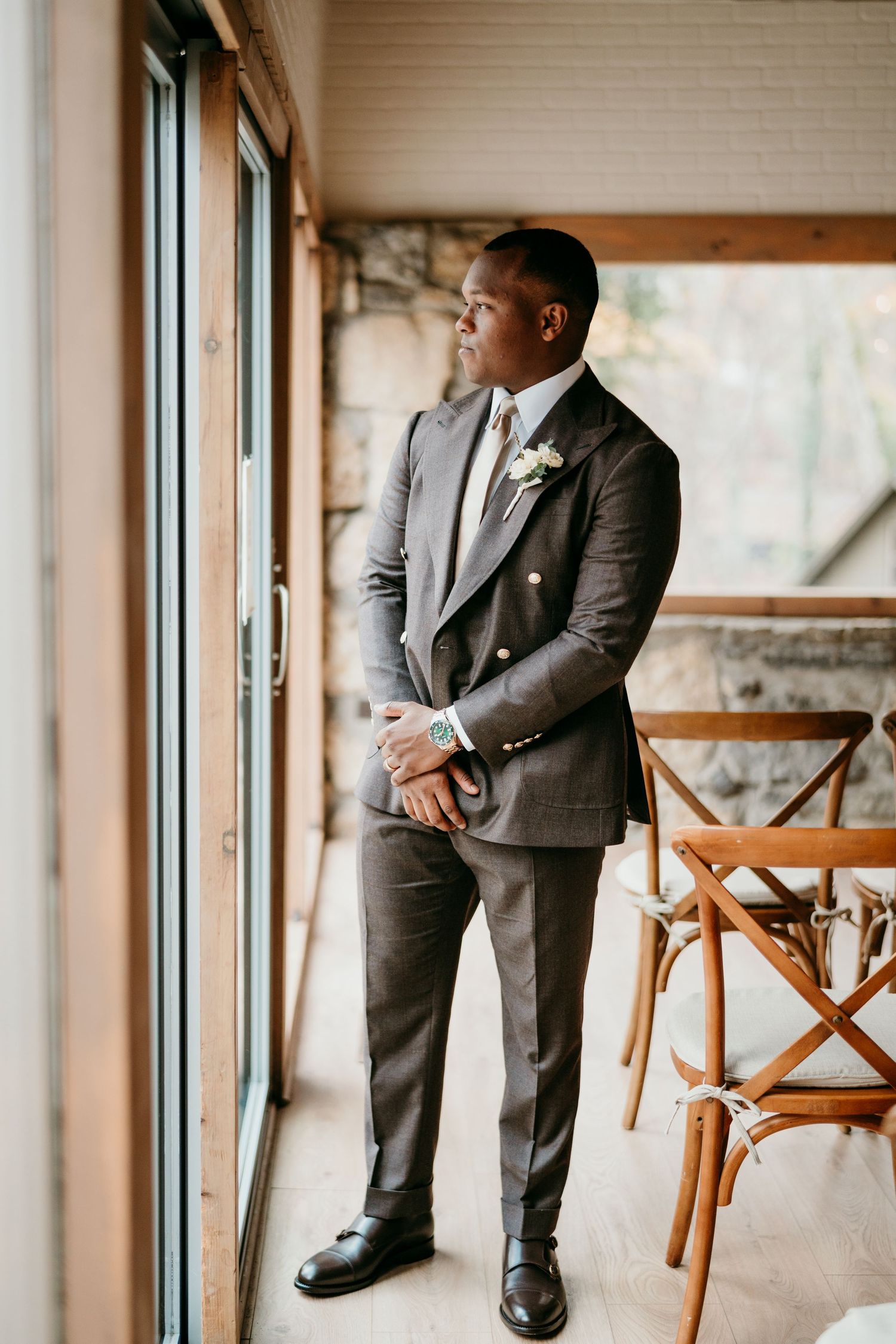 A groom in a charcoal grey suit and boutonniere looks thoughtfully out a window in an elegant indoor setting.