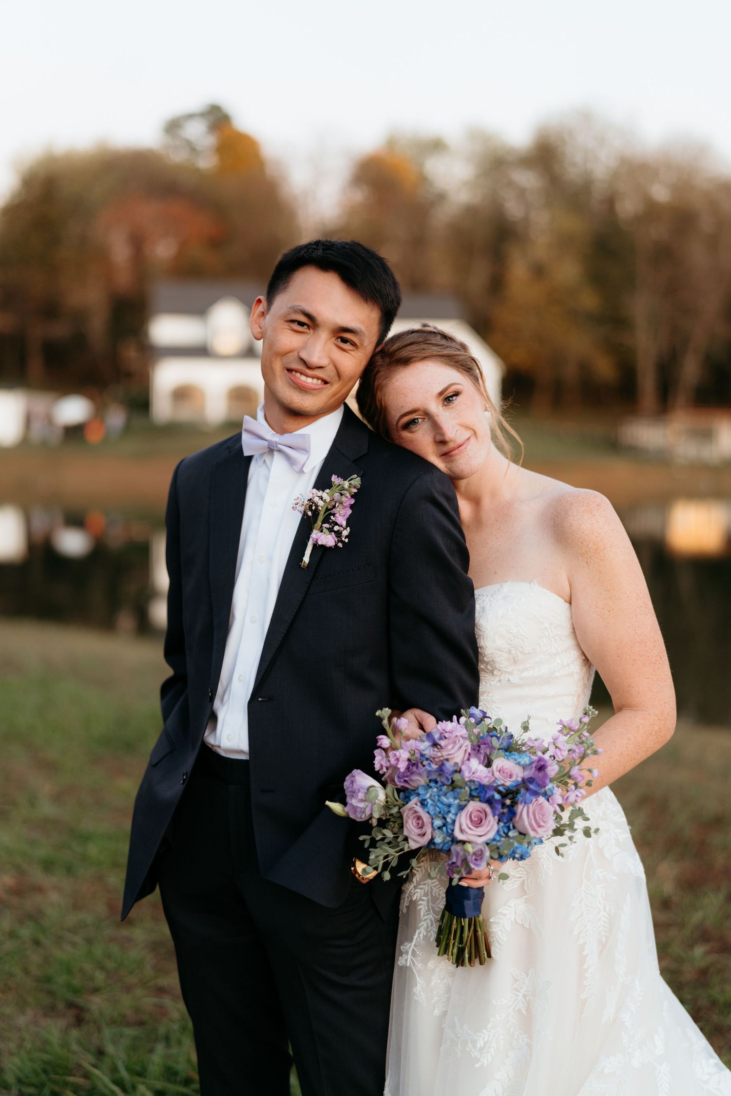 A couple in wedding attire poses outdoors with a purple and blue bouquet on a fall afternoon.