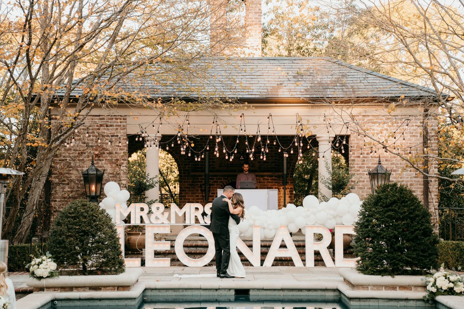 Rustic outdoor wedding venue with illuminated last name signs and twinkle lights under a covered pavilion at dusk.