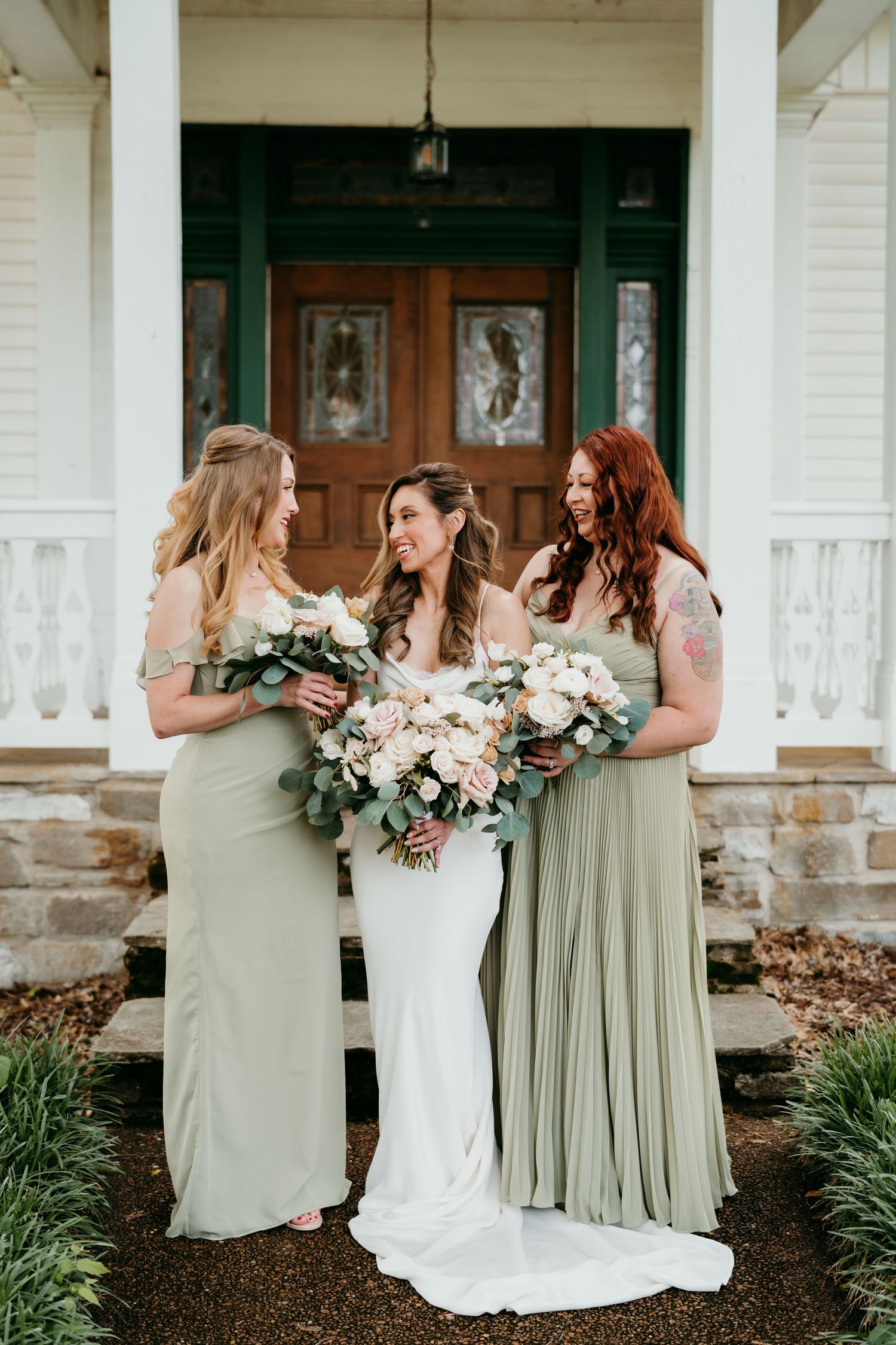sage green and white dresses hold floral bouquets while standing on a porch with an elegant green door at battle mountain farm