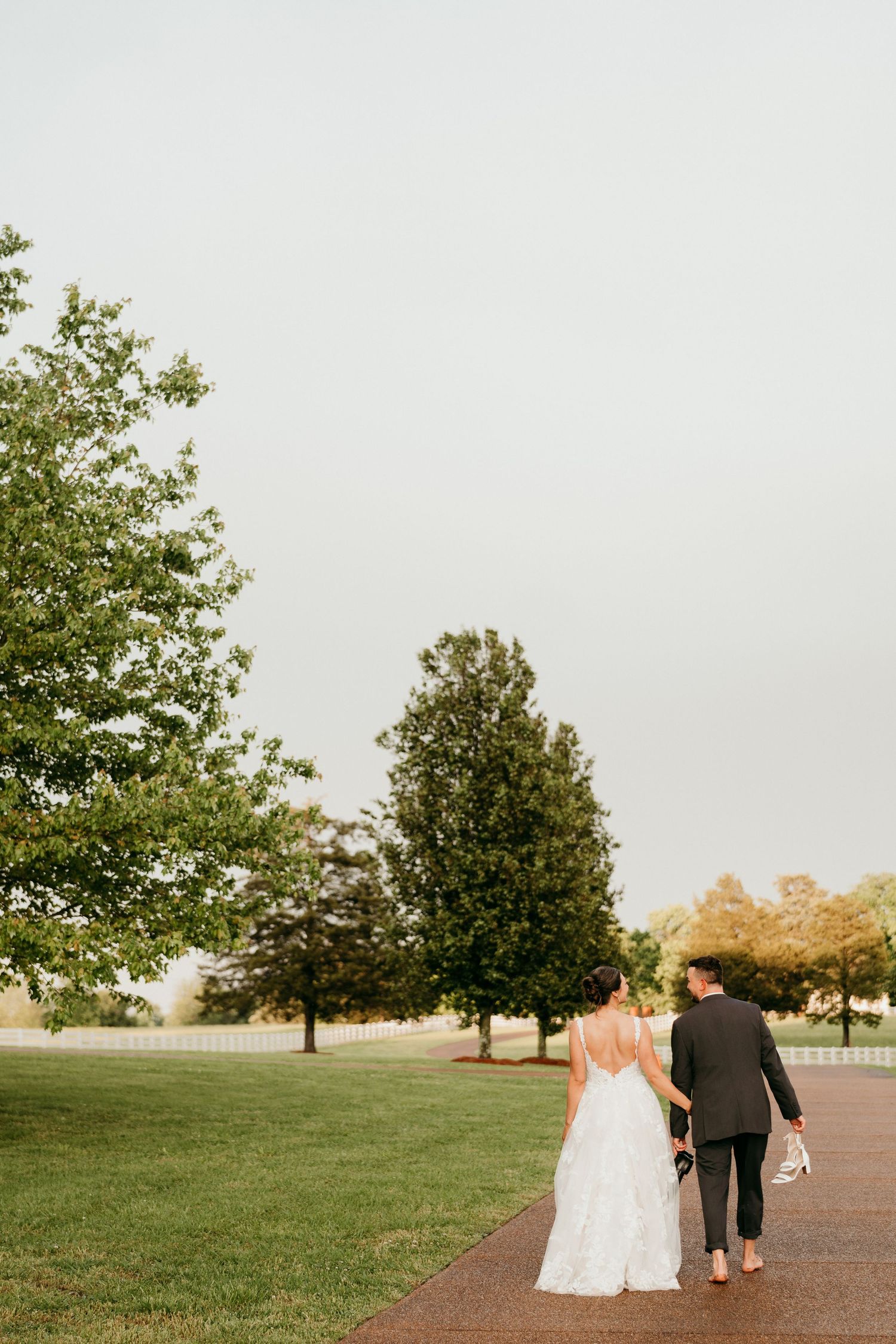 Couple walking barefoot on path surrounded by trees while holding their shoes after wedding ceremony.