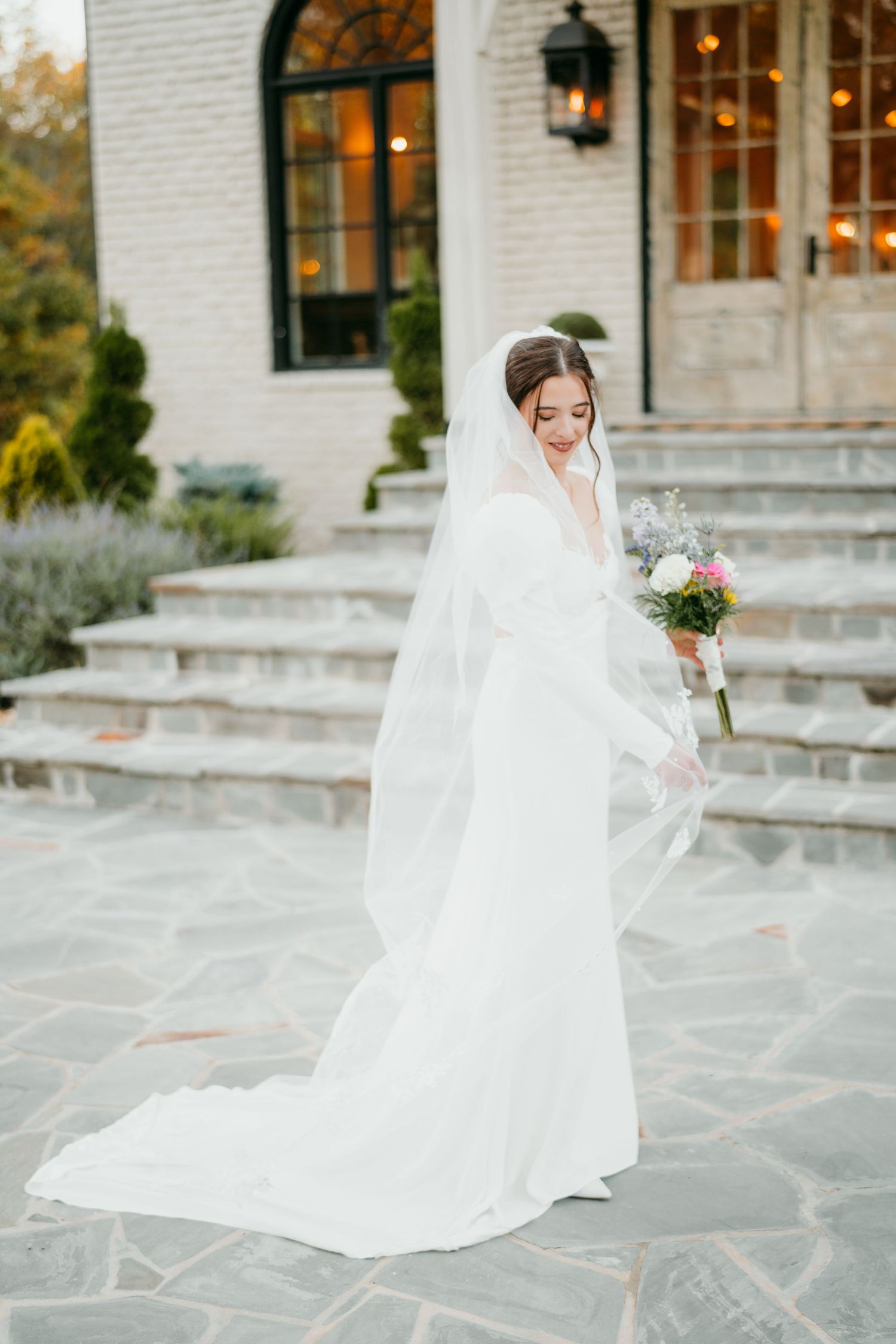 A bride in an elegant white wedding gown and flowing veil stands on stone steps outside a historic building with arched windows.