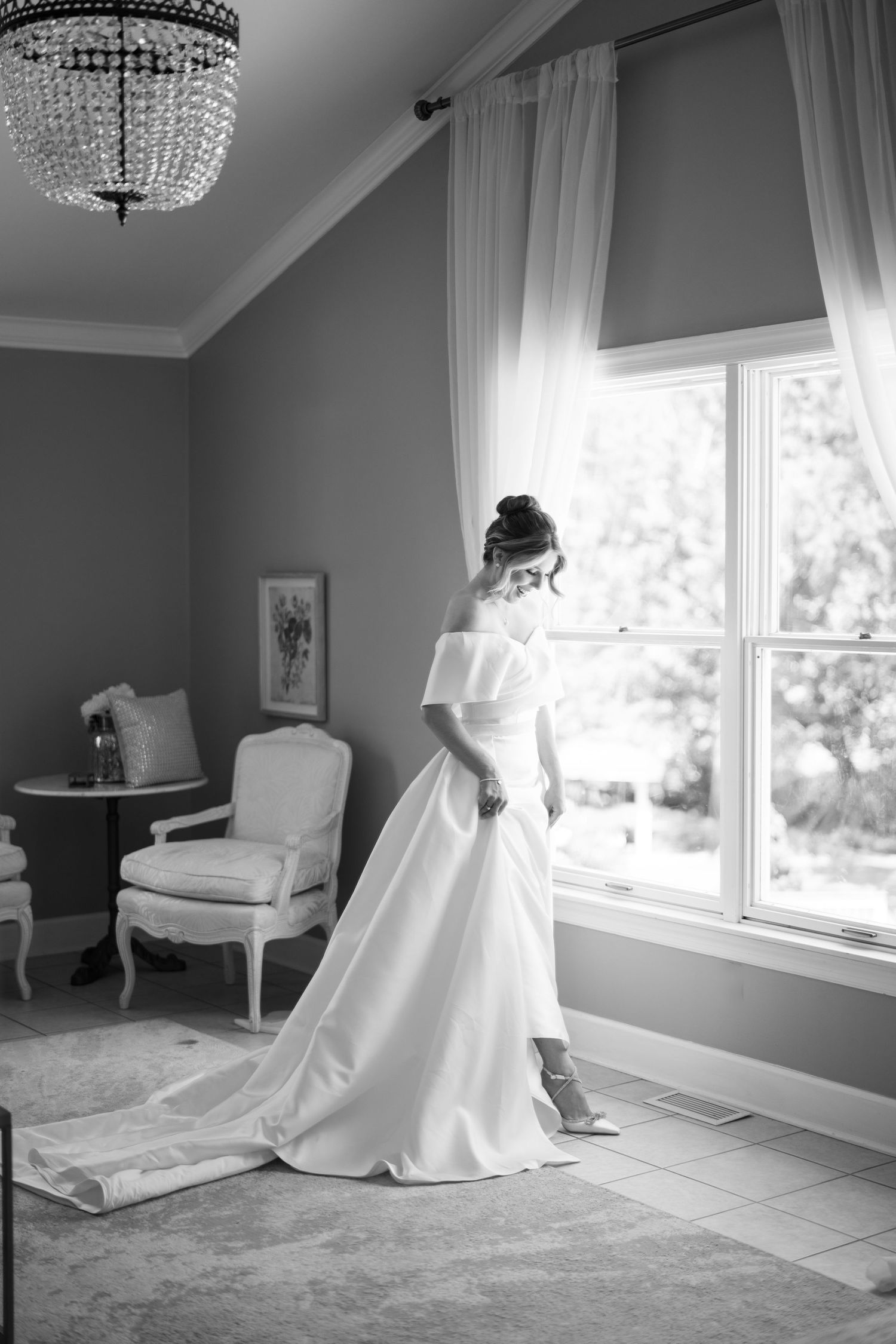 Elegant wedding dress silhouette against a window with crystal chandelier and vintage chair in black and white.