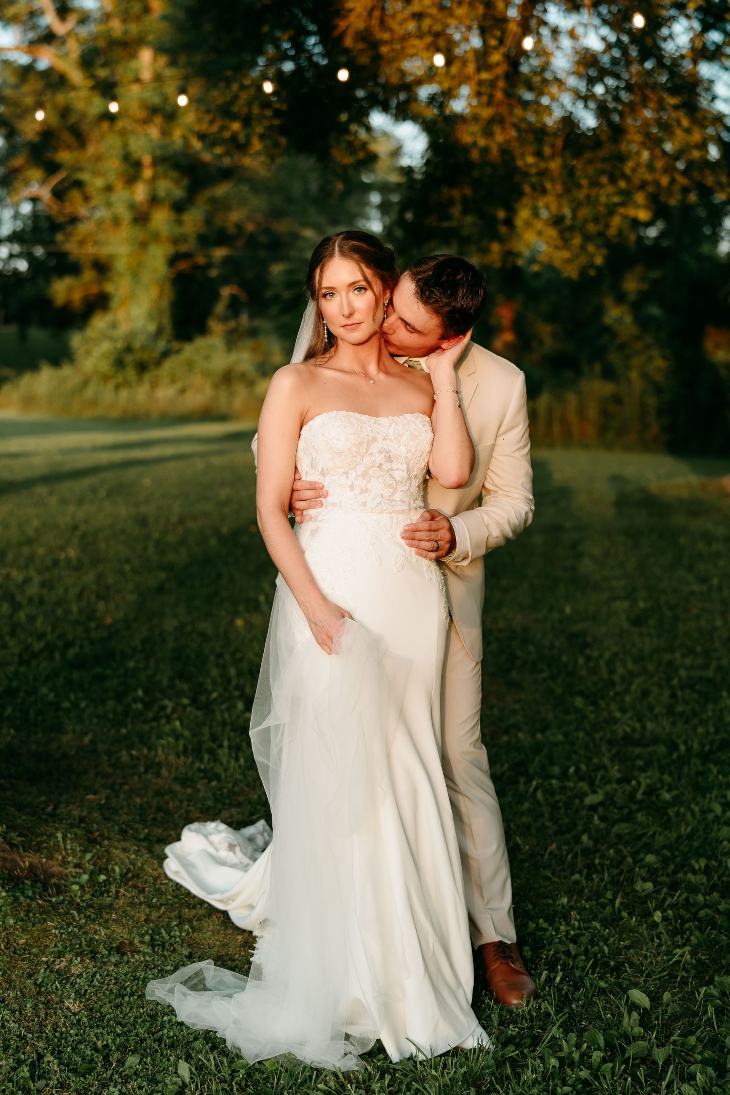 A romantic wedding couple embraces in a sunlit autumn field at golden hour as the sun sets behind fall foliage.