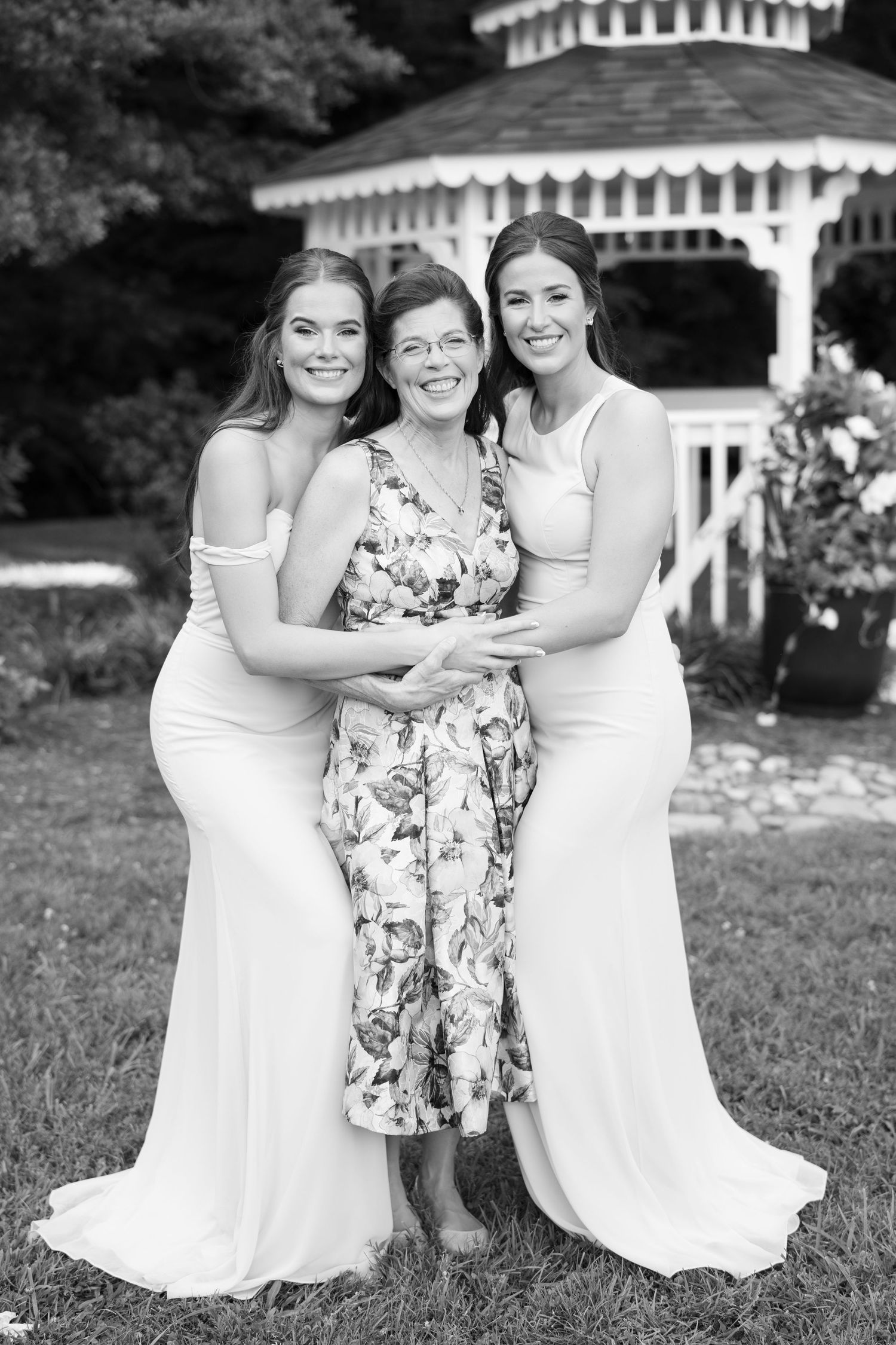 Three people in white dresses pose together in a garden setting with a white gazebo in the background.