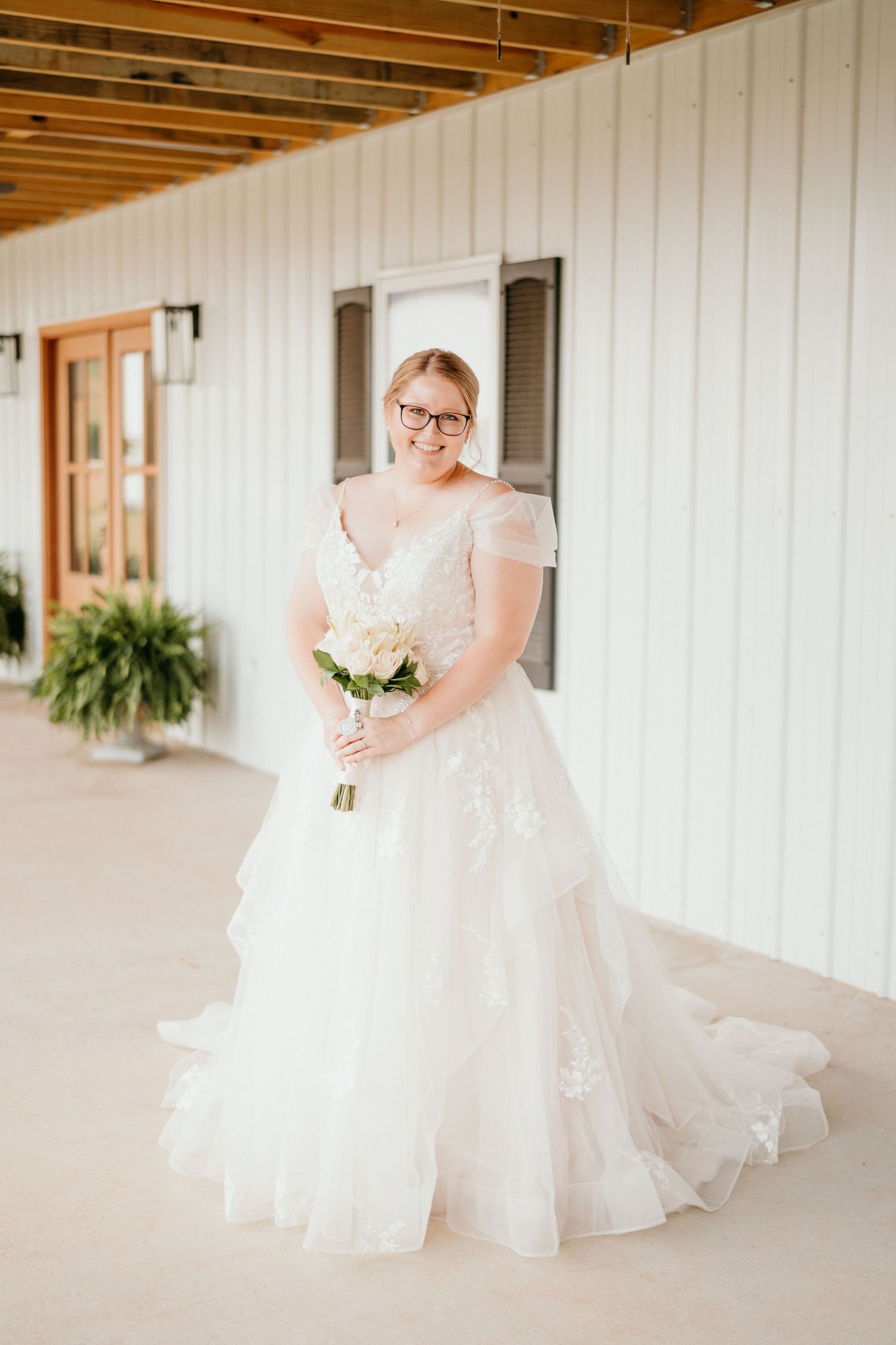 A beautiful wedding dress with sheer cap sleeves and tiered tulle skirt flows elegantly on a covered porch.