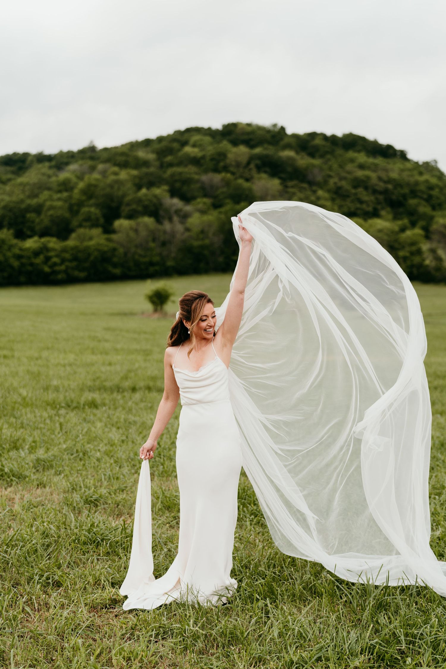 A bride in a flowing white dress stands in a green field as her veil billows dramatically in the wind.