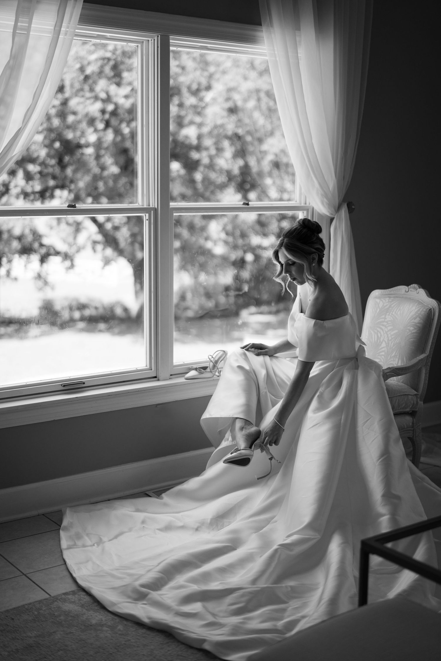 A black and white portrait of someone in a wedding dress sitting by a window.