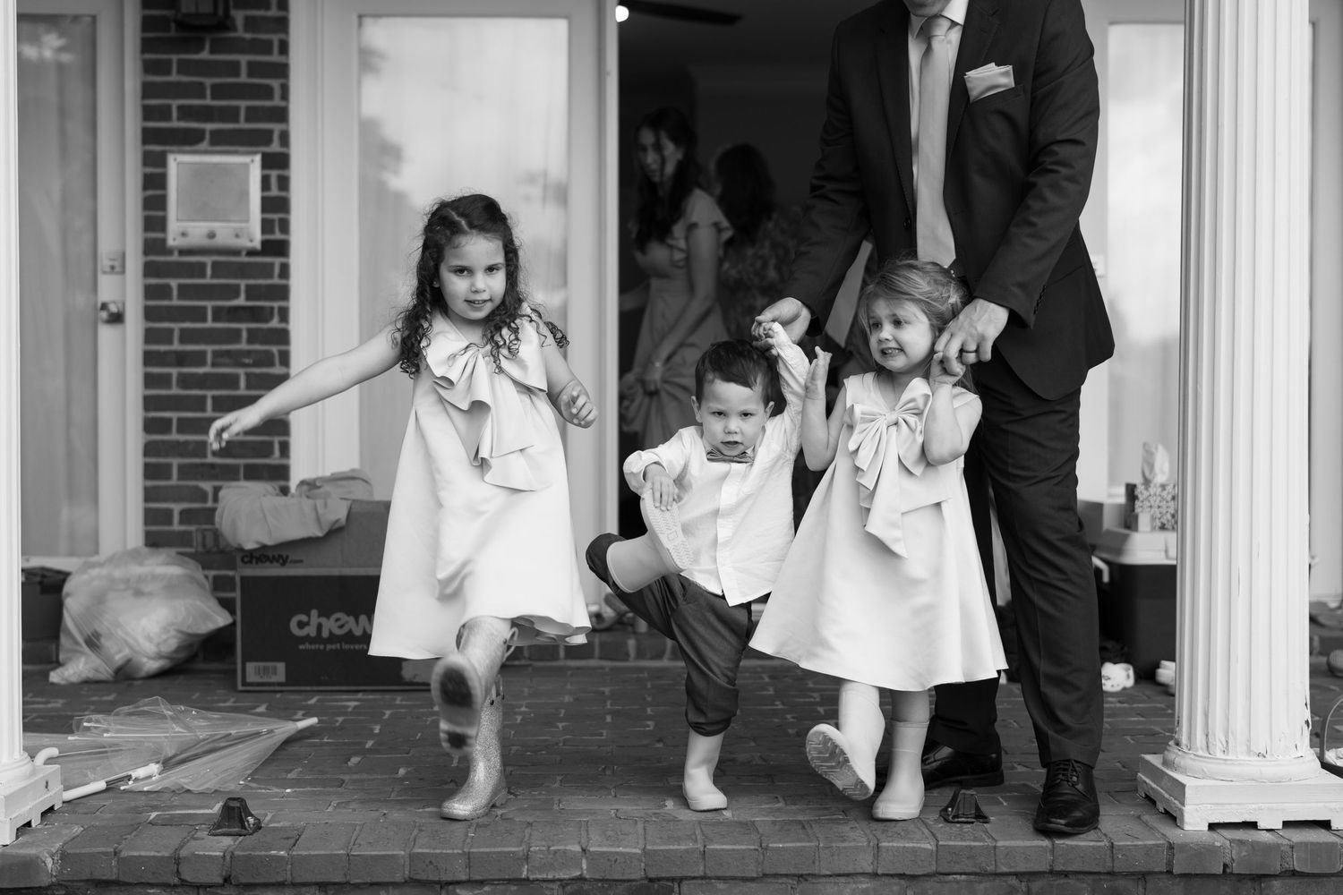 Three children in white dresses dance and twirl joyfully on the front porch of a brick building.