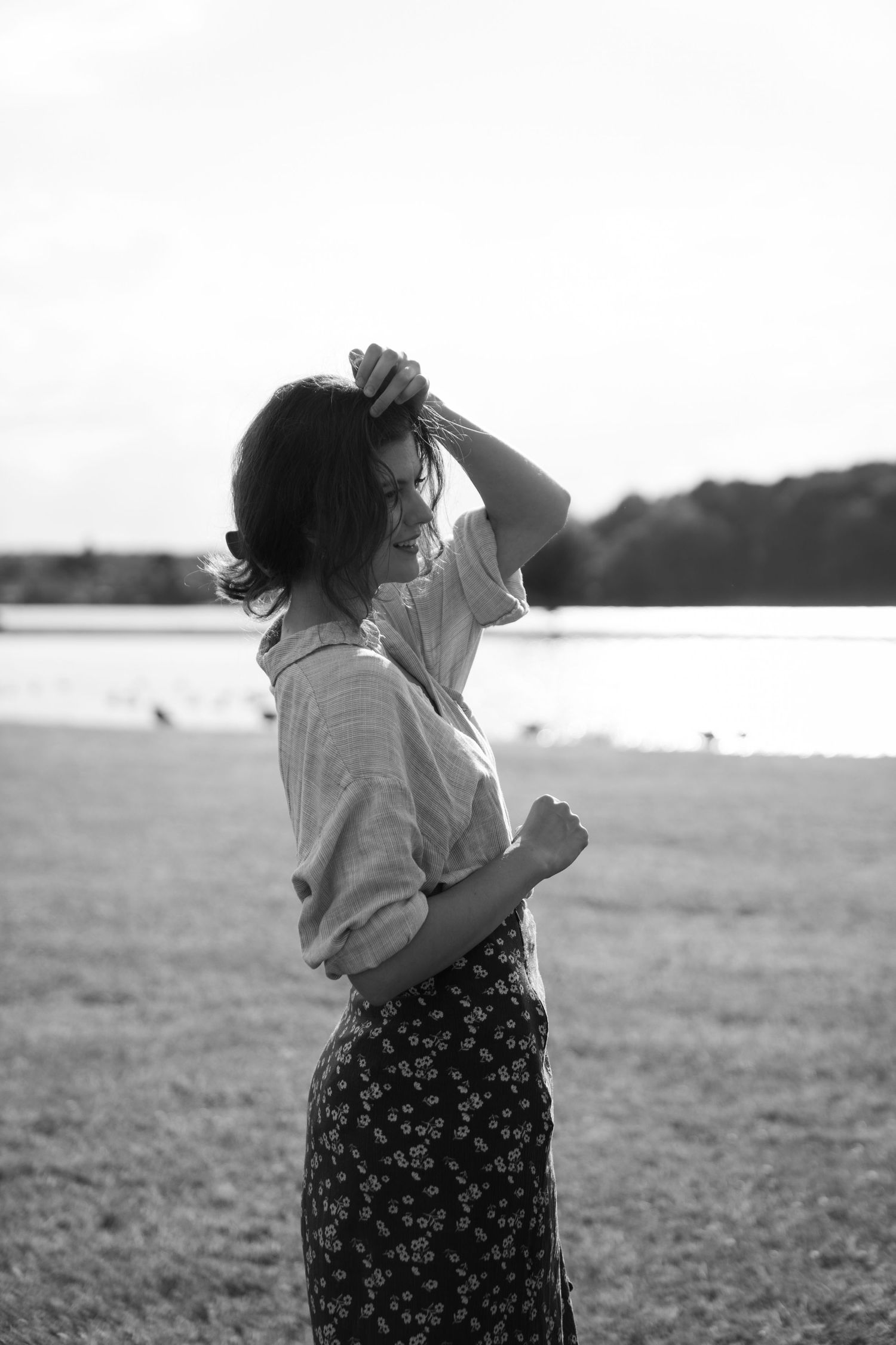 Black and white portrait of person in floral skirt by lakeside running fingers through hair.
