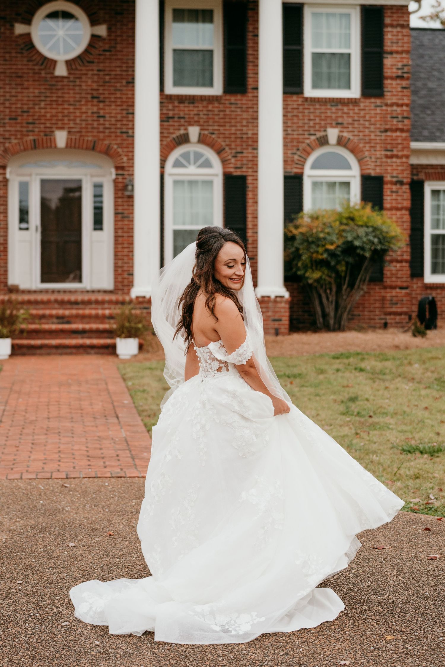 A beautiful bride in a flowing white wedding dress twirls gracefully in front of a classic brick home.