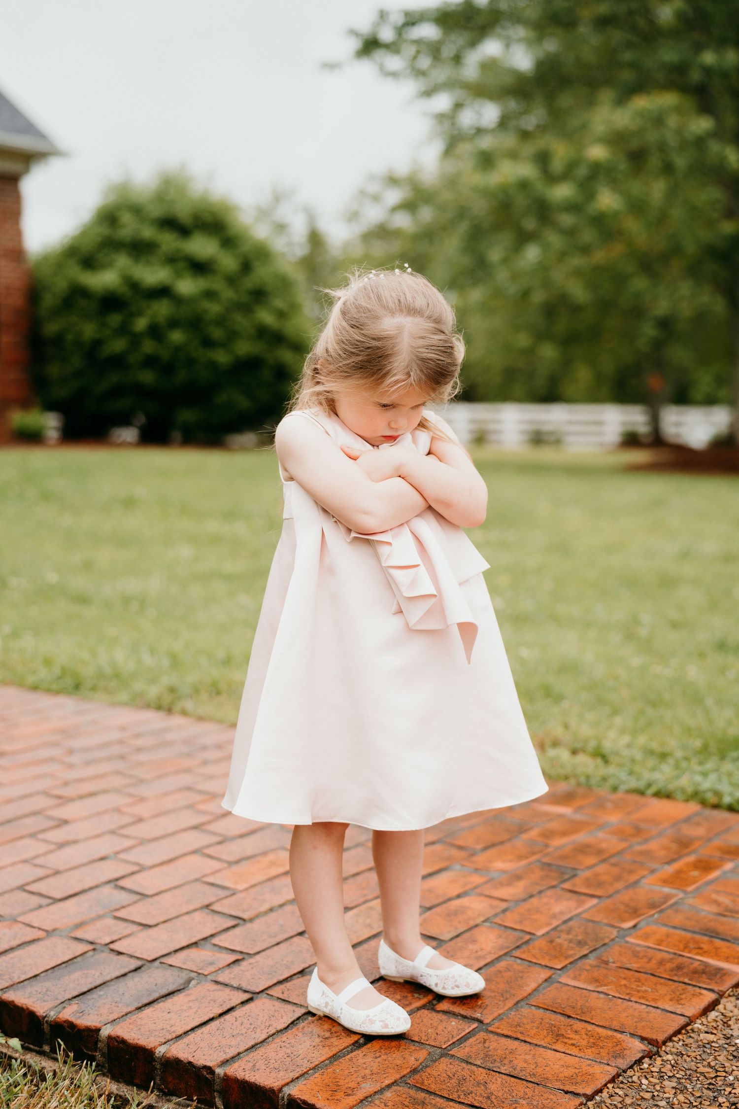 A small child in a white dress sits curled up on red brick pavement in a garden setting.