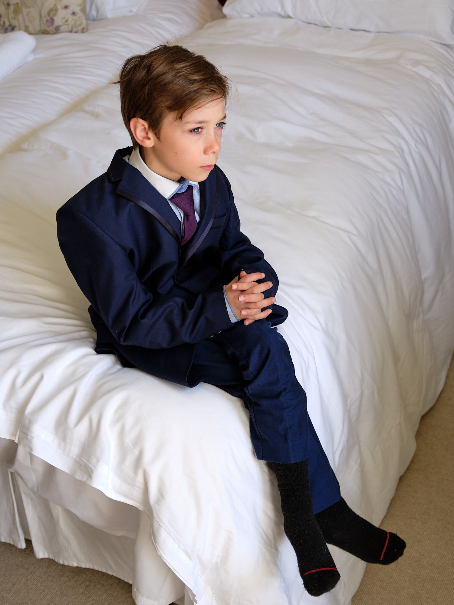 Young attendant in navy suit sits thoughtfully on white bedding during wedding preparations in Brighton, East Sussex.