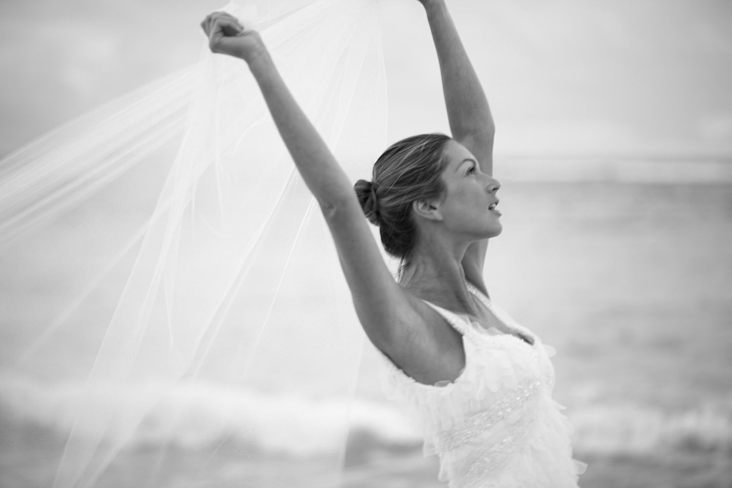 Black and white photo of bride in wedding dress with arms raised joyfully against sky.