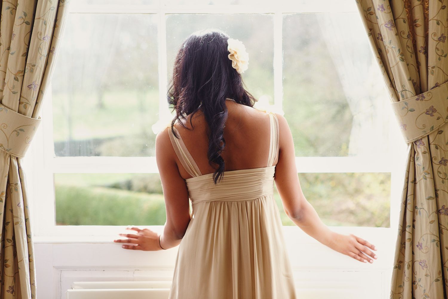 Row of bridesmaids in cream colored dresses standing by large windows.