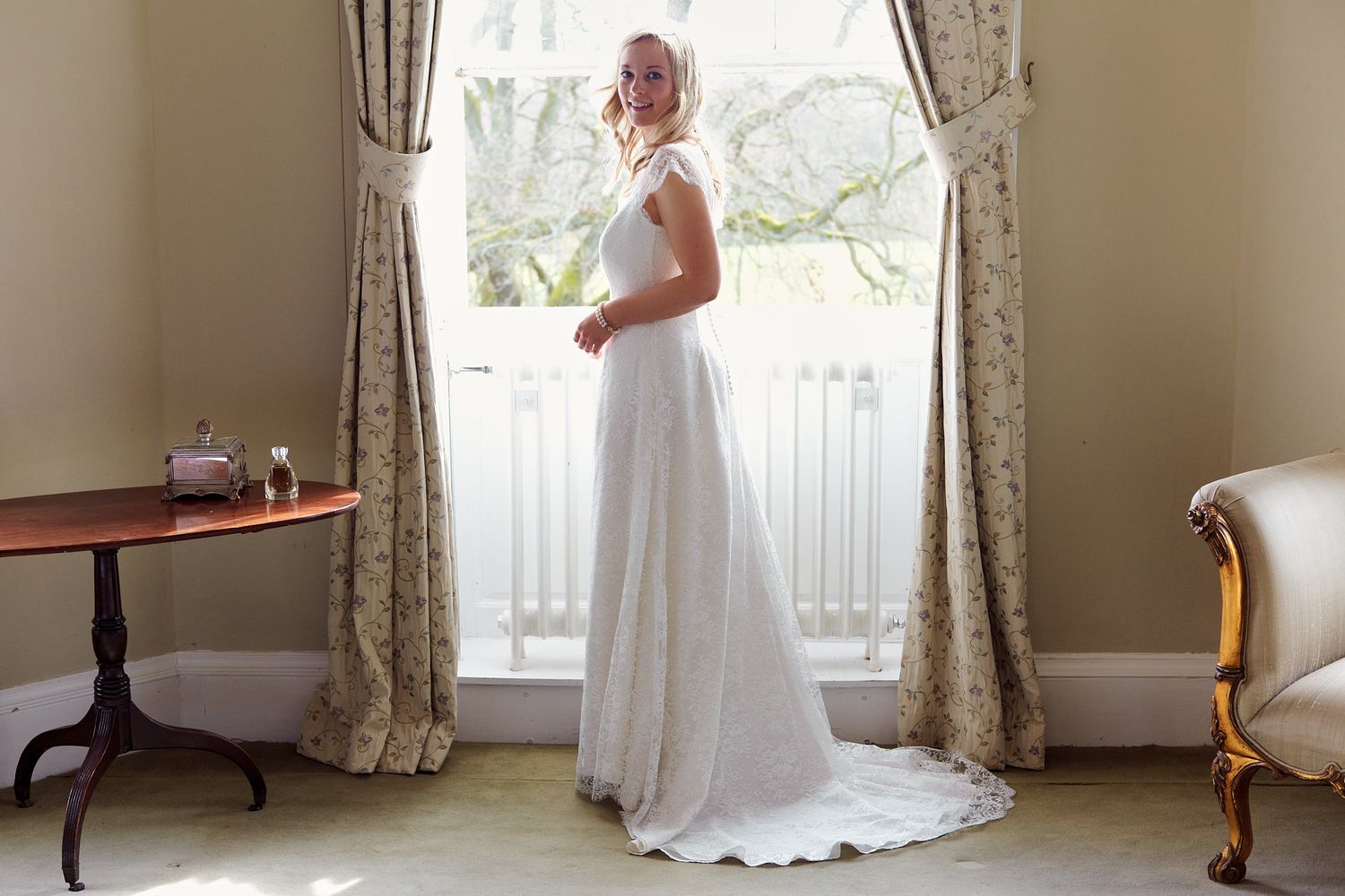 Wedding dress silhouette against window light in elegant room with drapes.