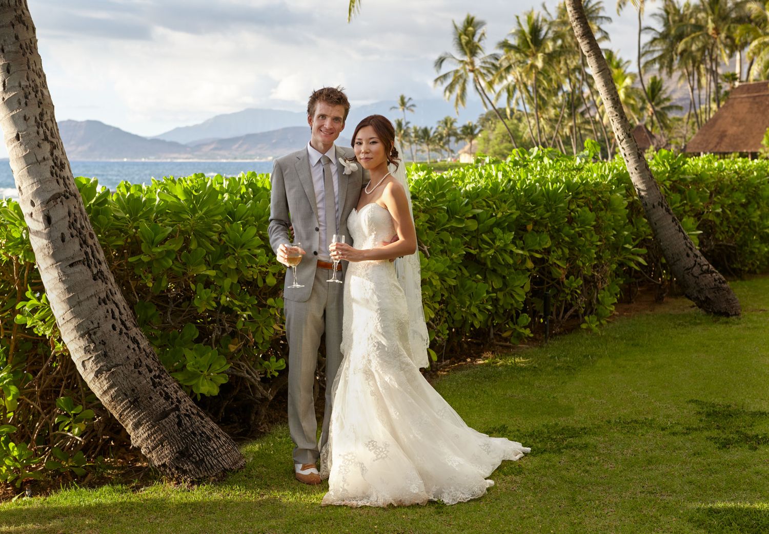 Newlyweds pose on tropical lawn with palm trees and ocean view in background at destination wedding in Hawaii.
