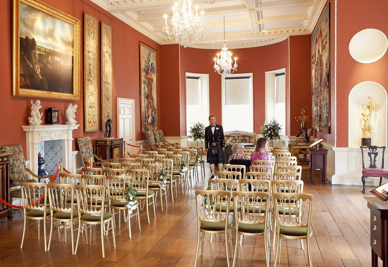 Elegant red-walled ballroom with crystal chandeliers and gold chiavari chairs set for wedding ceremony.