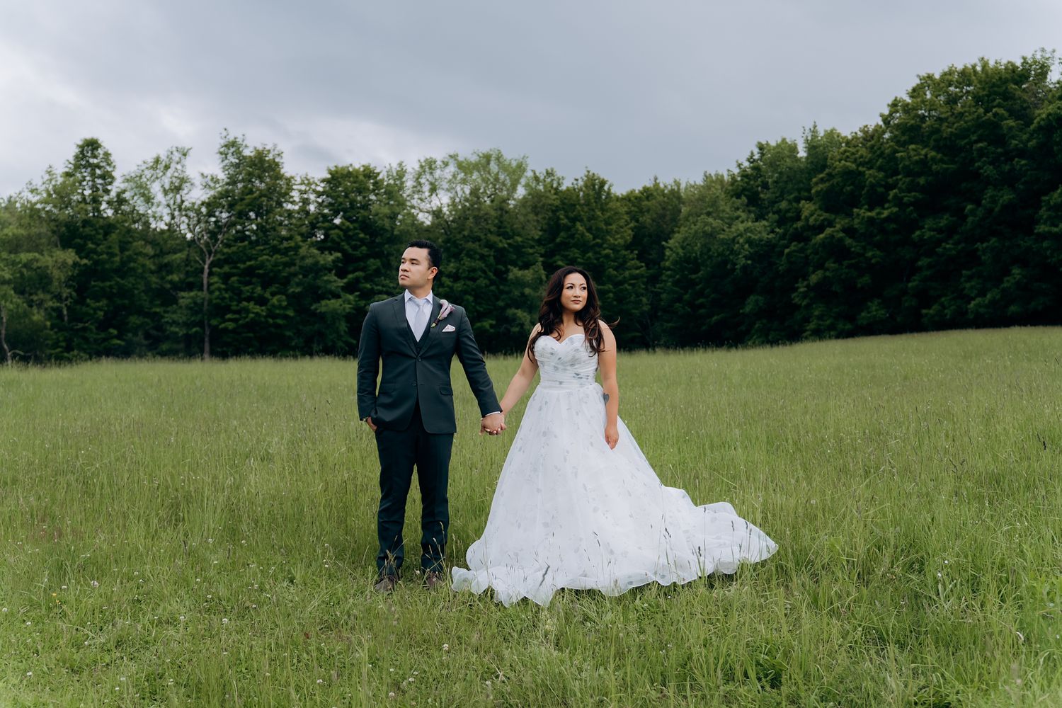 Bride and groom holding hands in a vast green meadow with a dramatic cloudy sky backdrop.