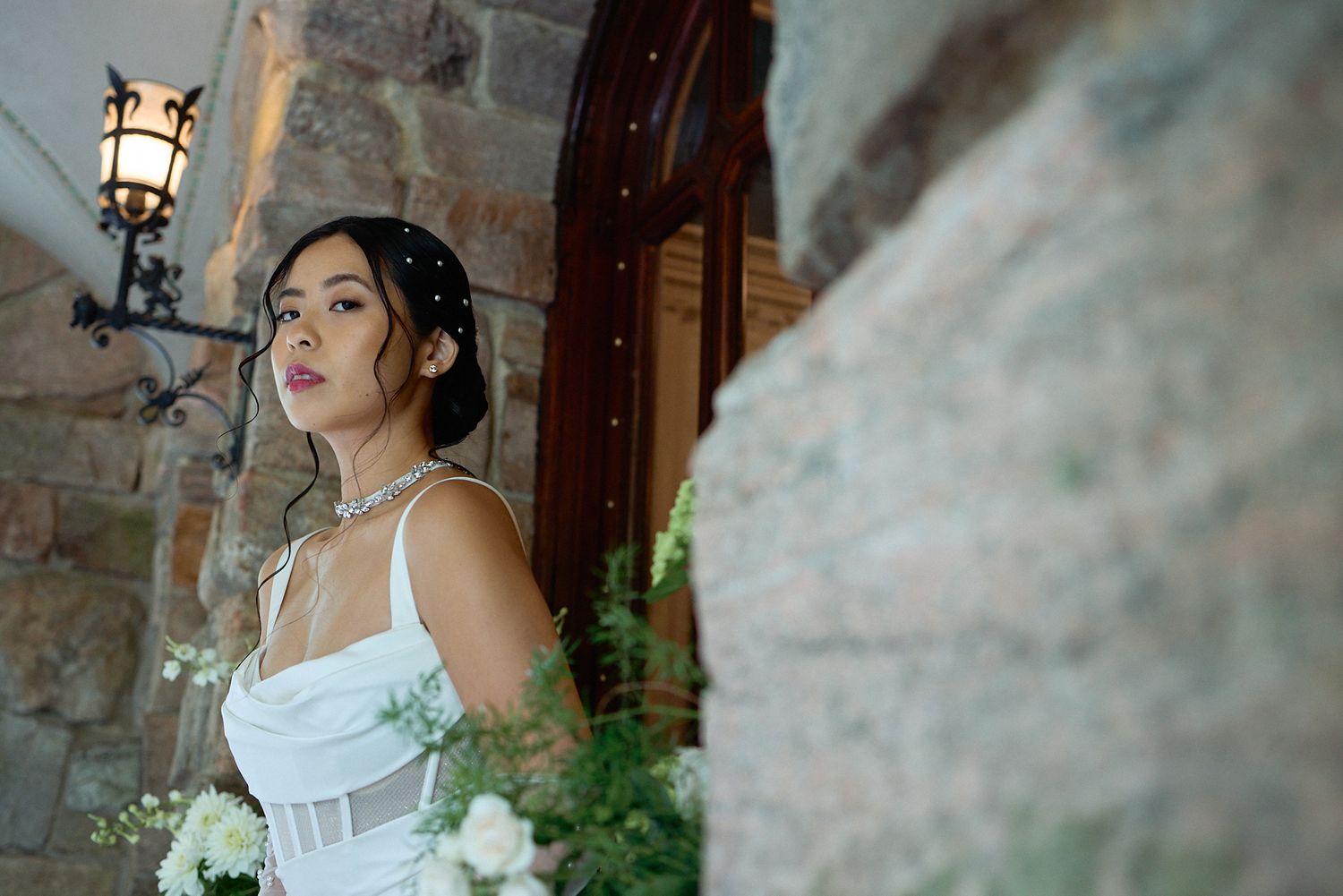 Stunning bride in white corset wedding gown gazes over shoulder beside stone wall with floral arrangements.