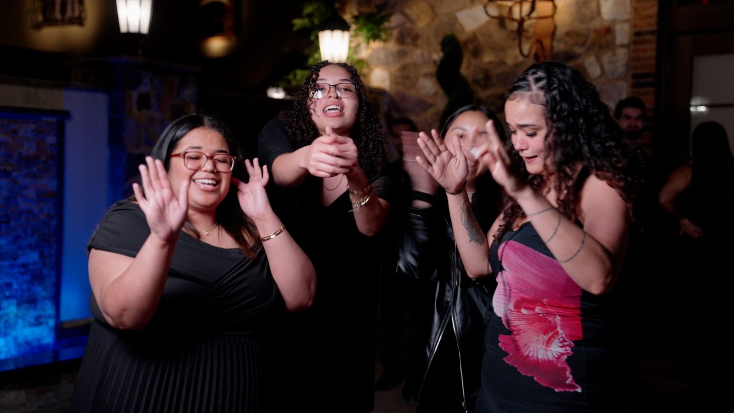 Four women laughing and clapping together in a warmly lit restaurant or event venue.
