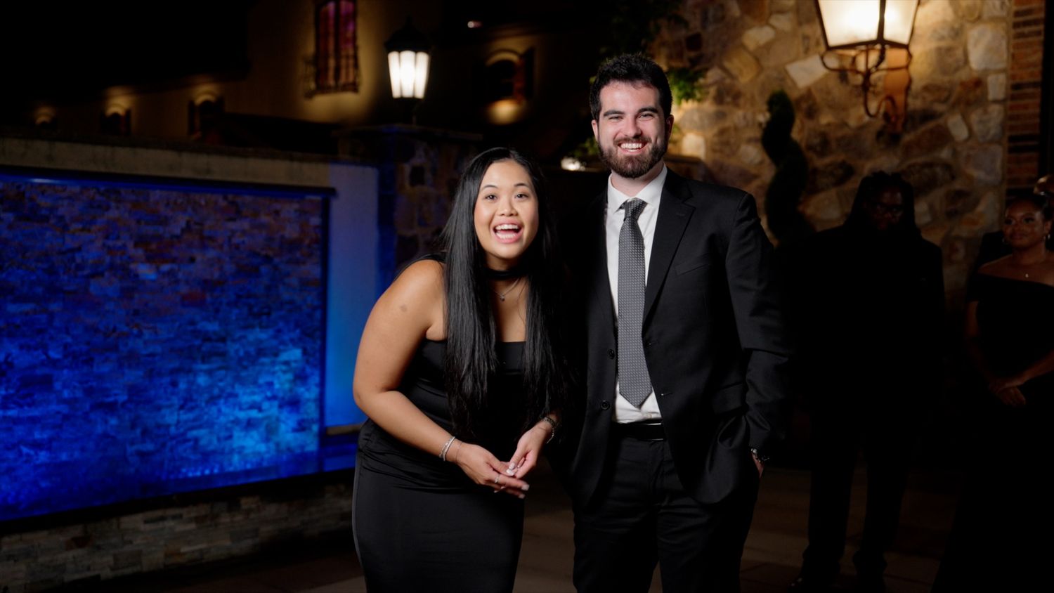 Smiling woman in black dress posing with a man in a suit at a nighttime event.