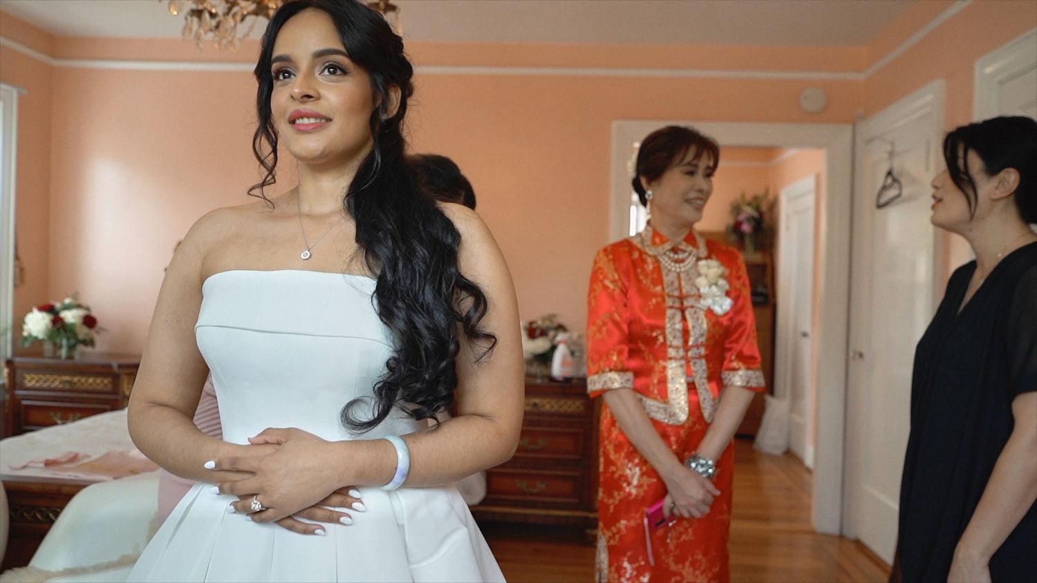 Bride in white strapless wedding dress standing with guests wearing orange traditional clothing.