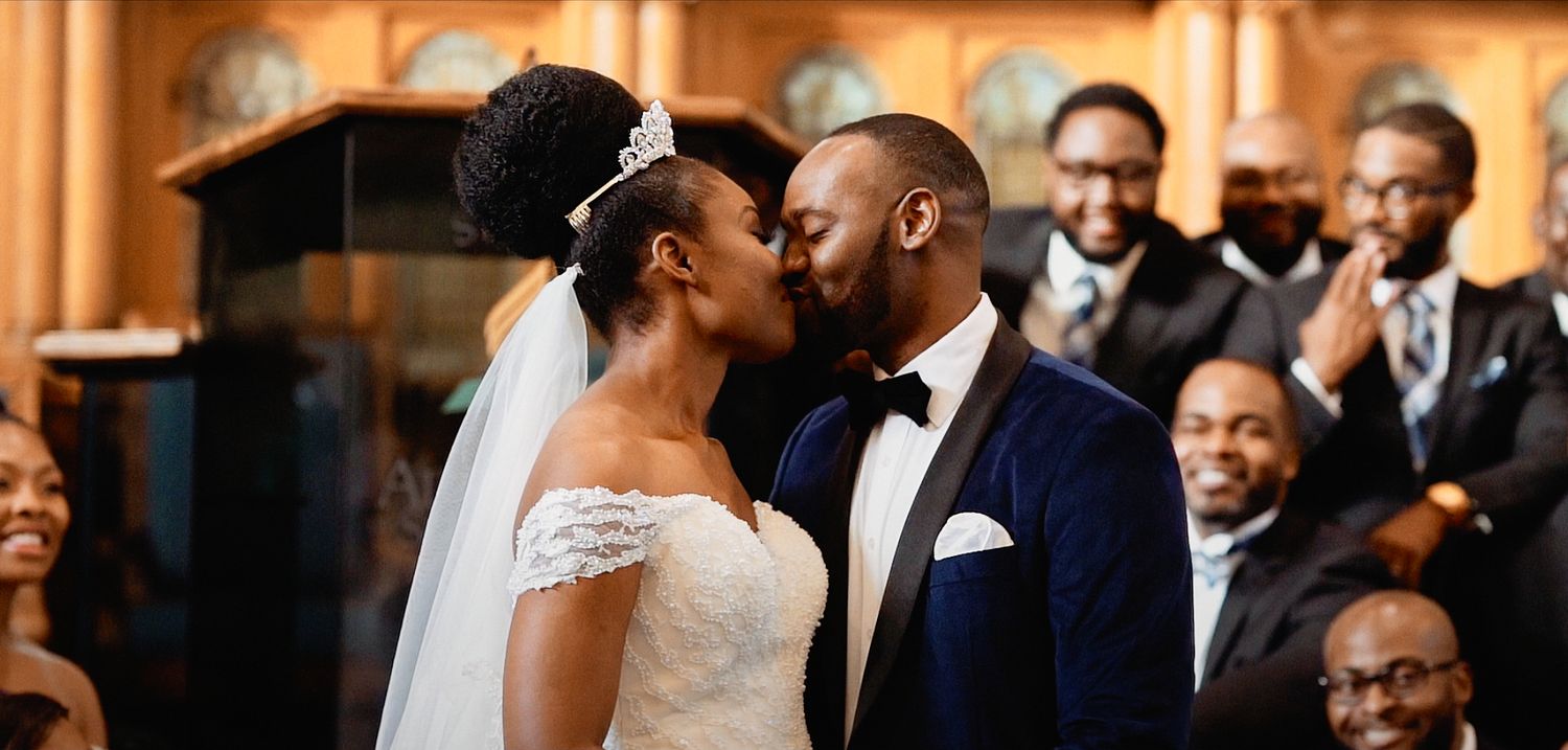 Black couple sharing first kiss at wedding altar with joyful guests in background.