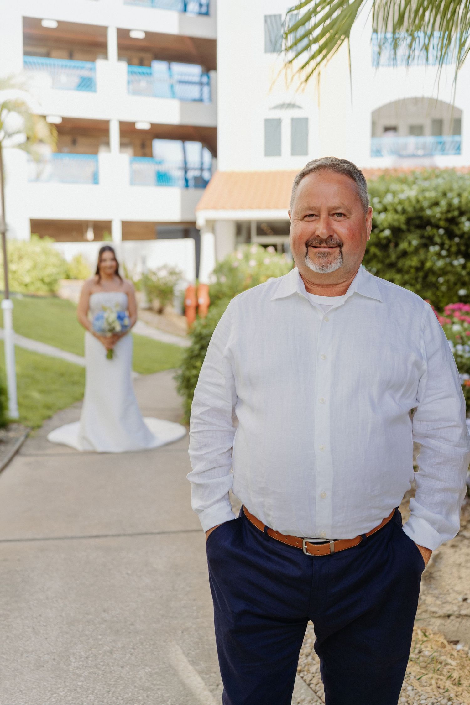 Groom in white shirt smiles outdoors with bride in white gown holding blue bouquet in background near resort.