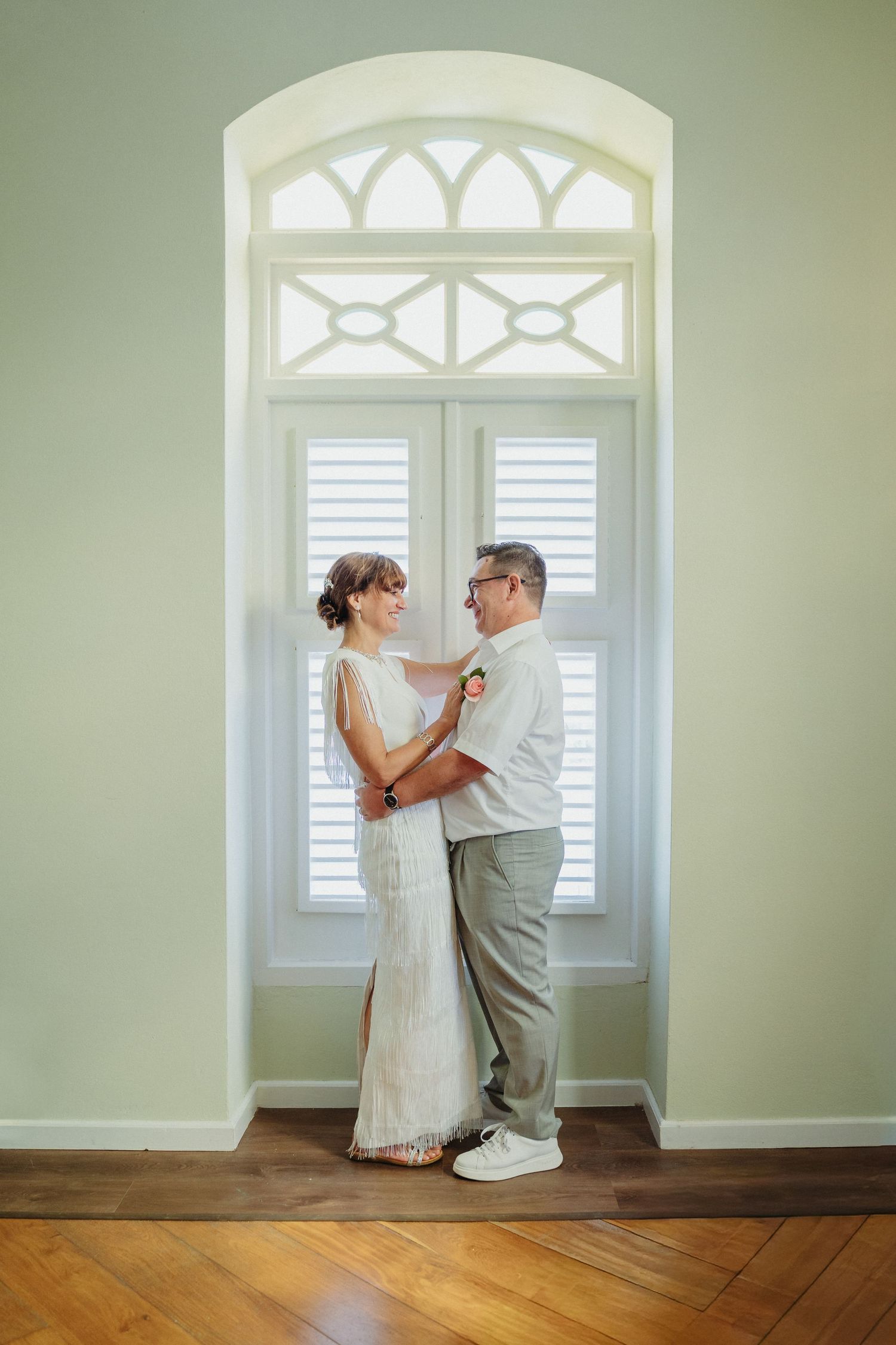 A couple embraces by a vintage window with decorative arched details and white plantation shutters.