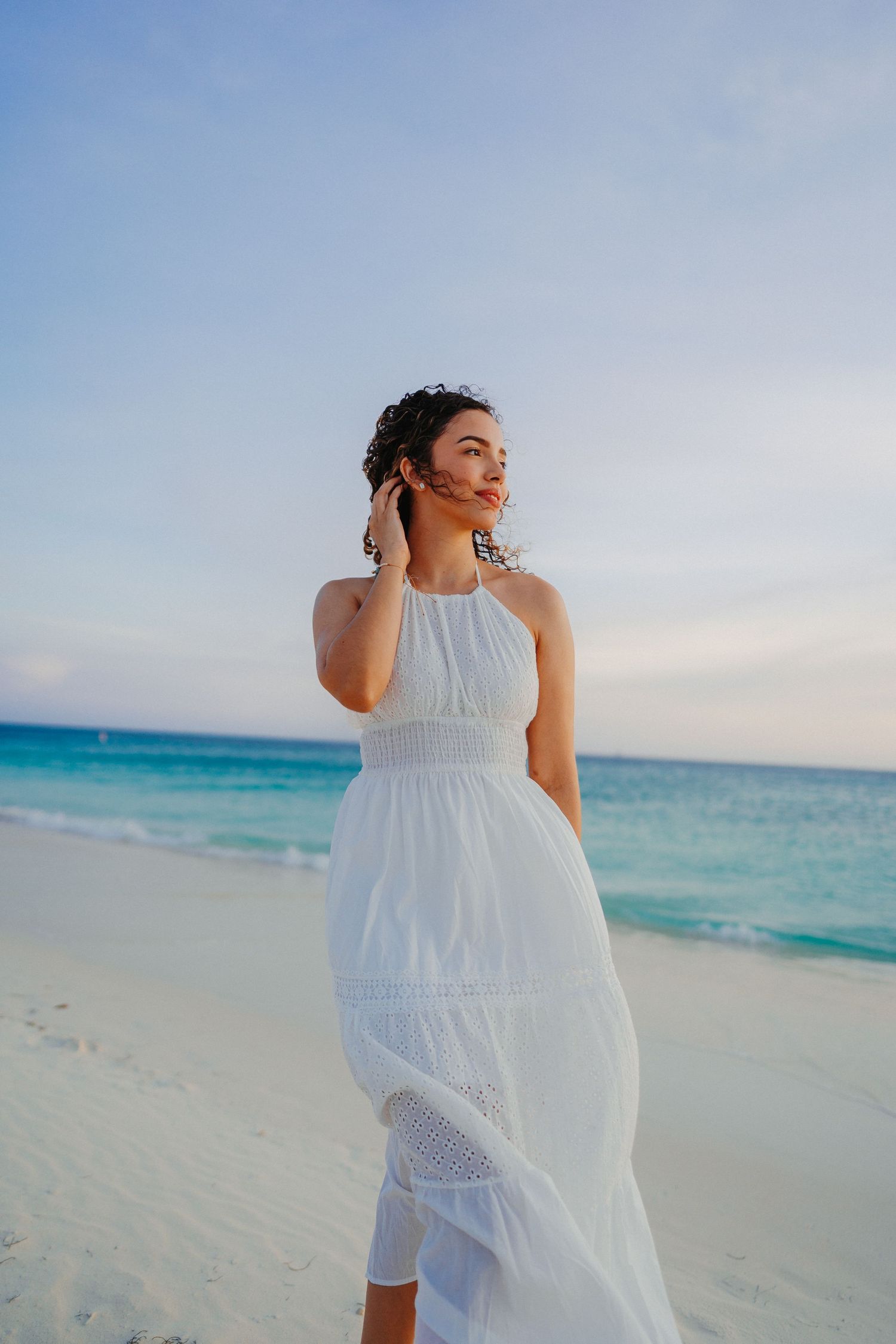 A person in a flowing white dress walks gracefully along a pristine beach with turquoise waters and clear skies.