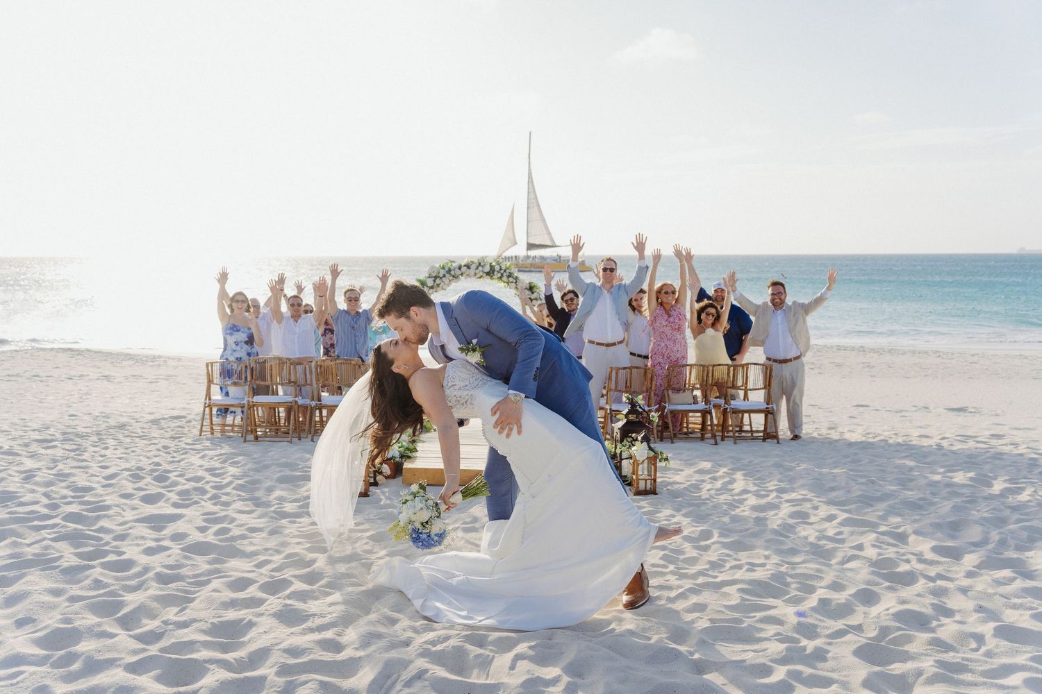Bride and groom share a romantic dip kiss on a white sand beach with cheering wedding guests behind them.