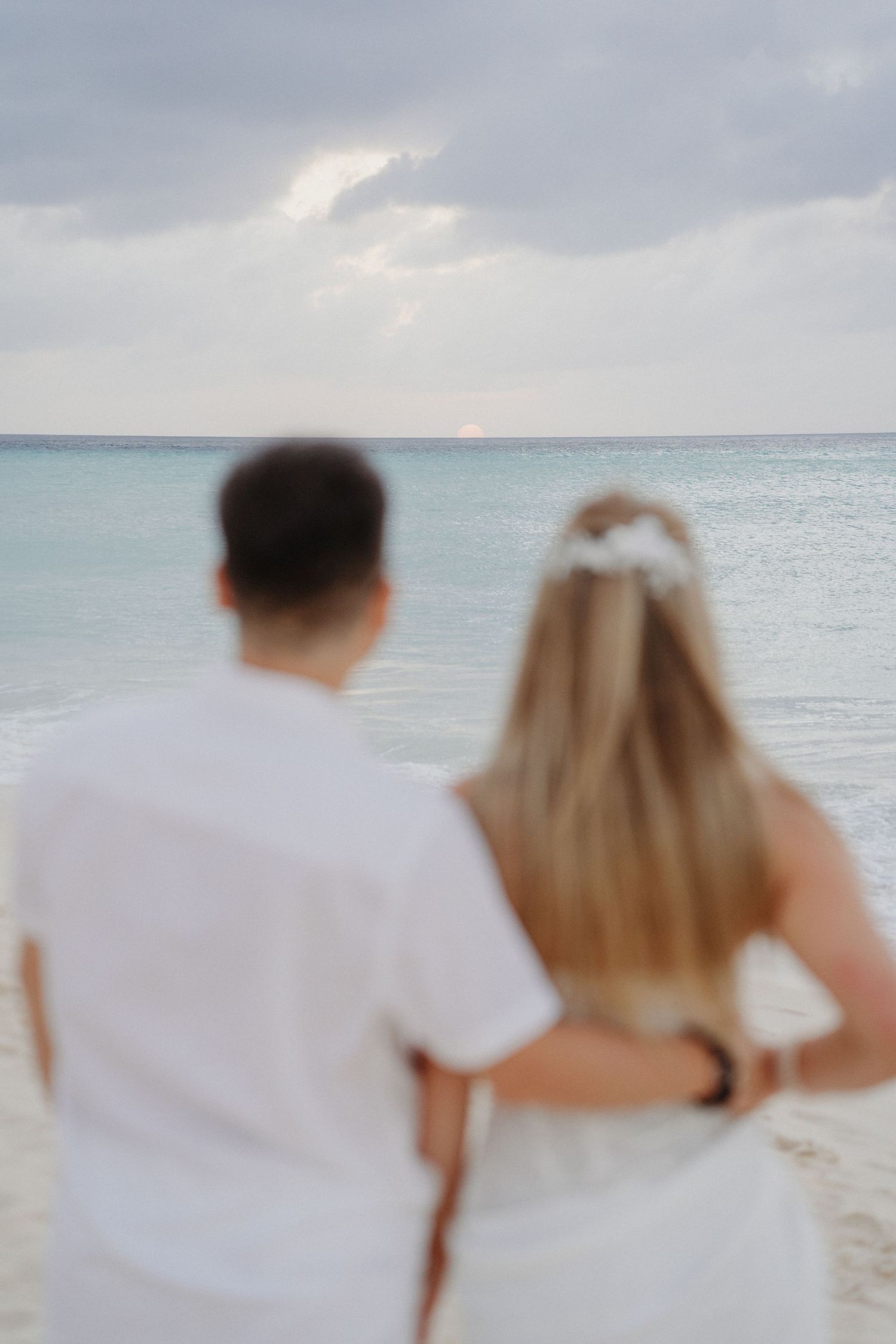 A couple in white attire walks together along a serene beach with cloudy skies and turquoise waters in the background.