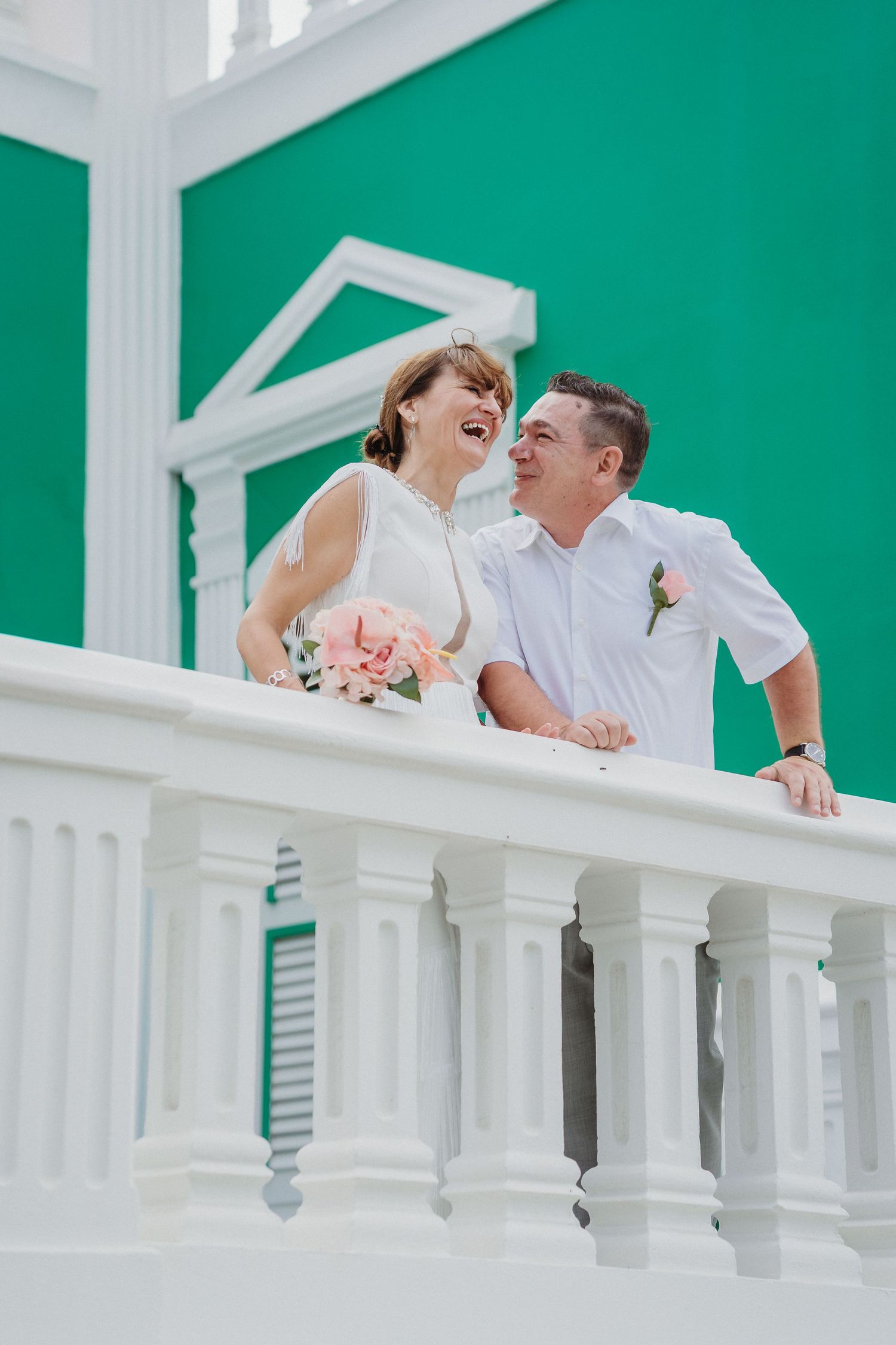 A romantic wedding couple shares an intimate moment on a white balcony with turquoise walls in the background.