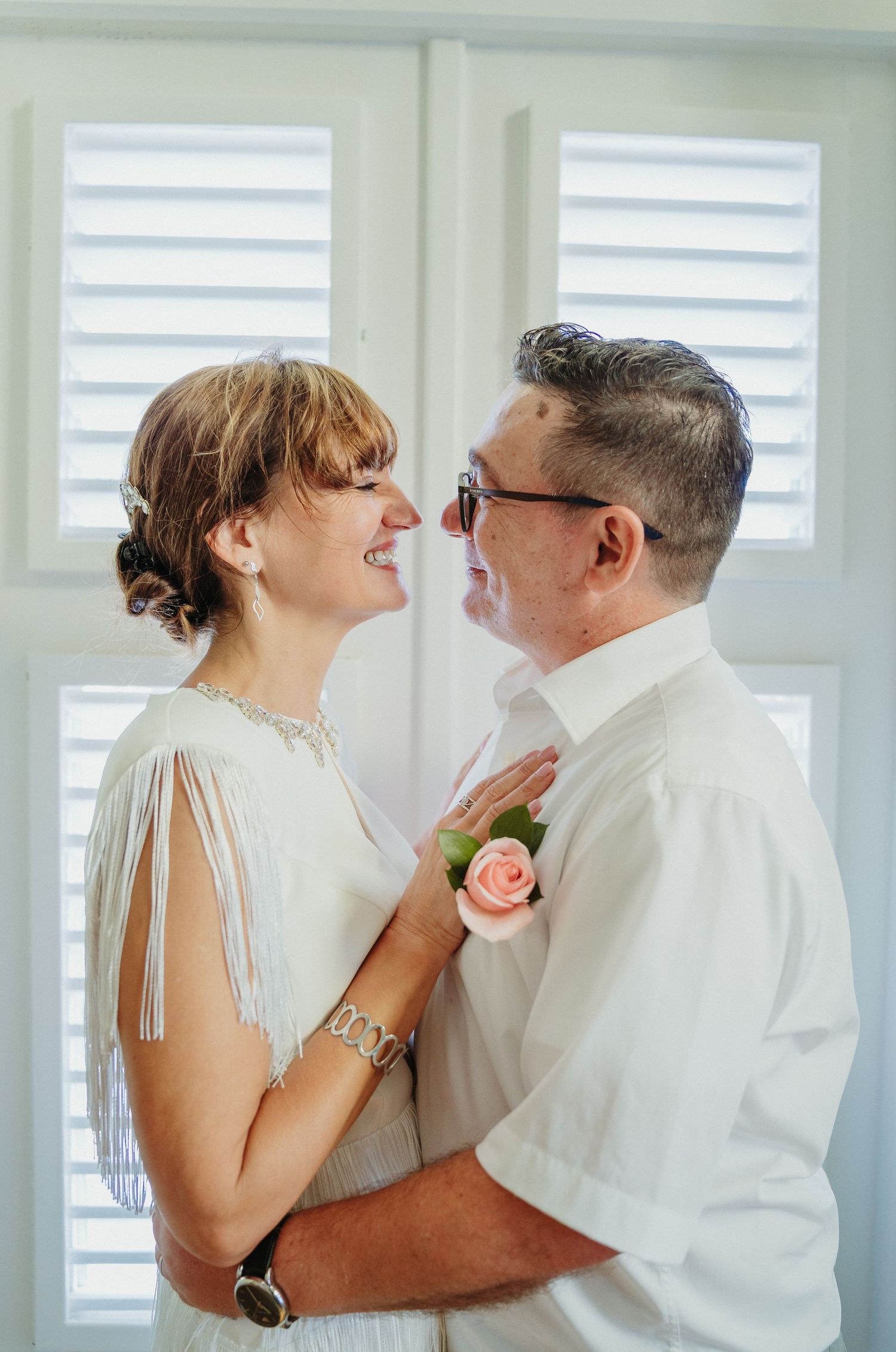 An intimate wedding moment captured indoors with white shuttered windows in the background.