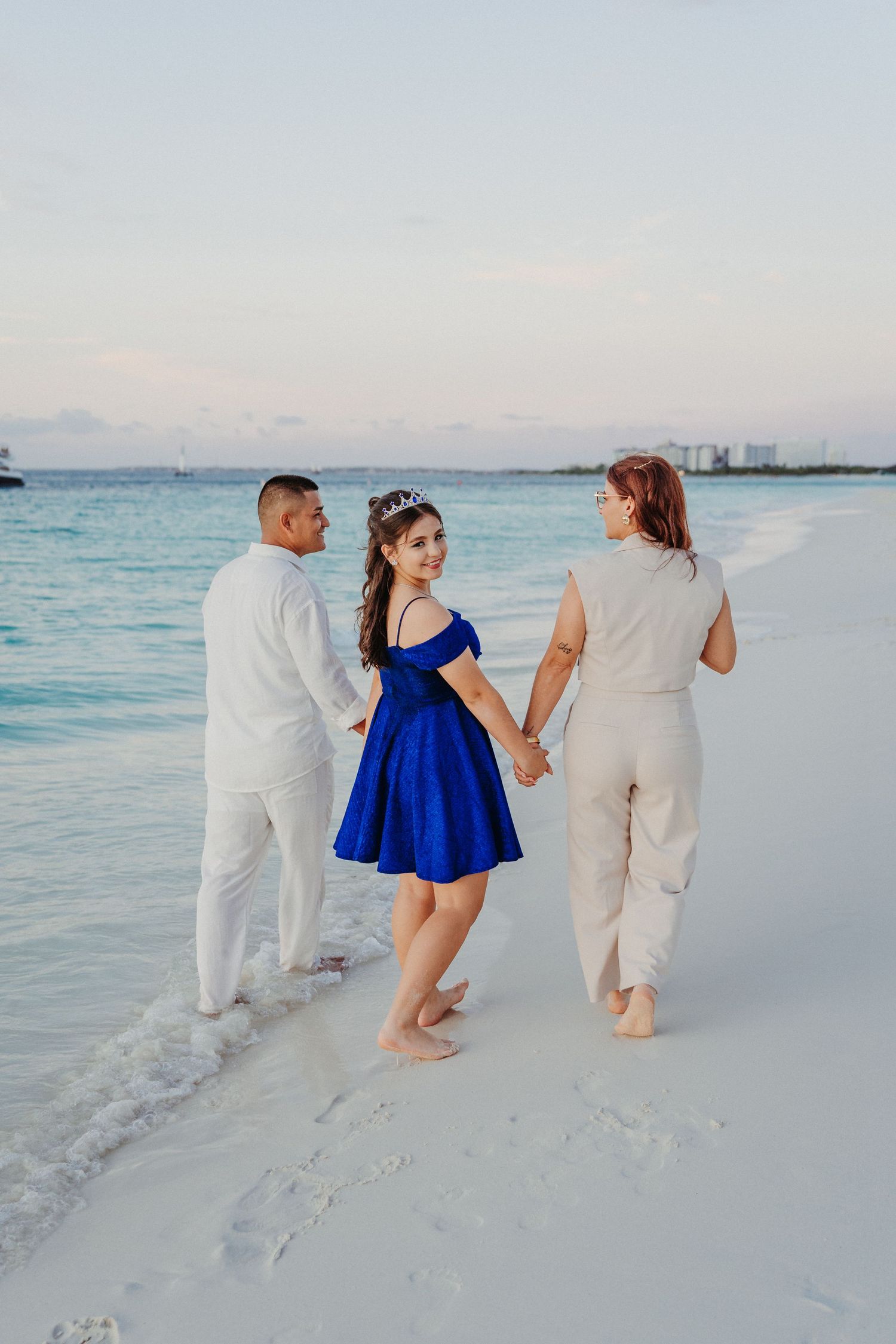 Three people in white clothing and blue dress walking along a pristine beach shoreline at sunset.