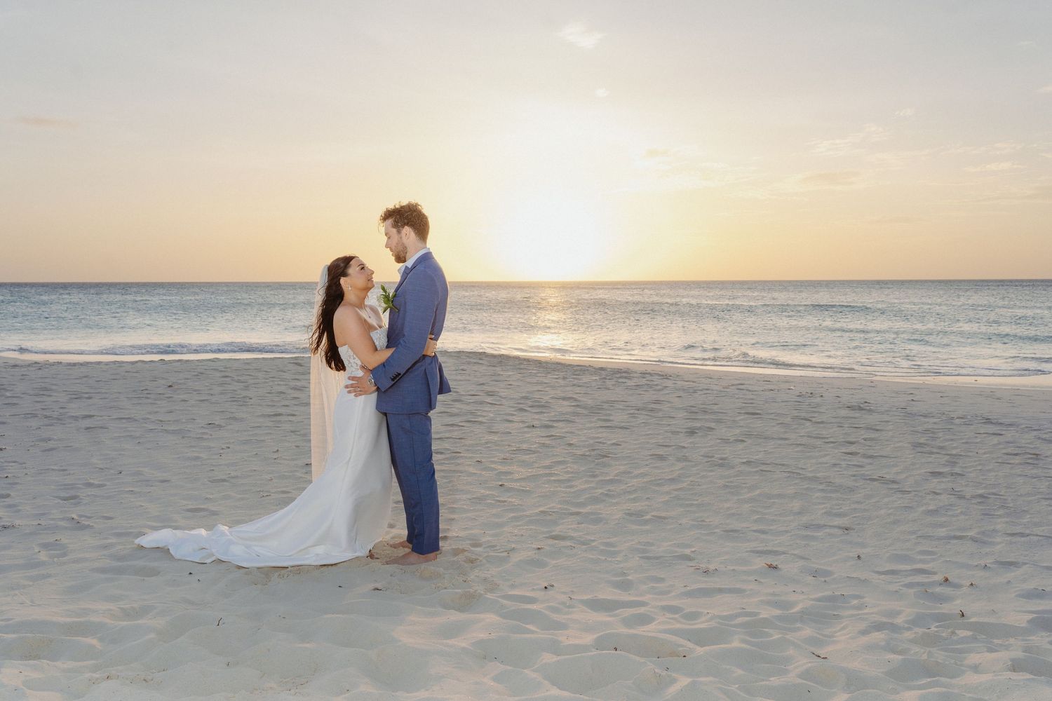 Bride and groom share a romantic moment on a sandy beach at sunset with ocean waves in the background.
