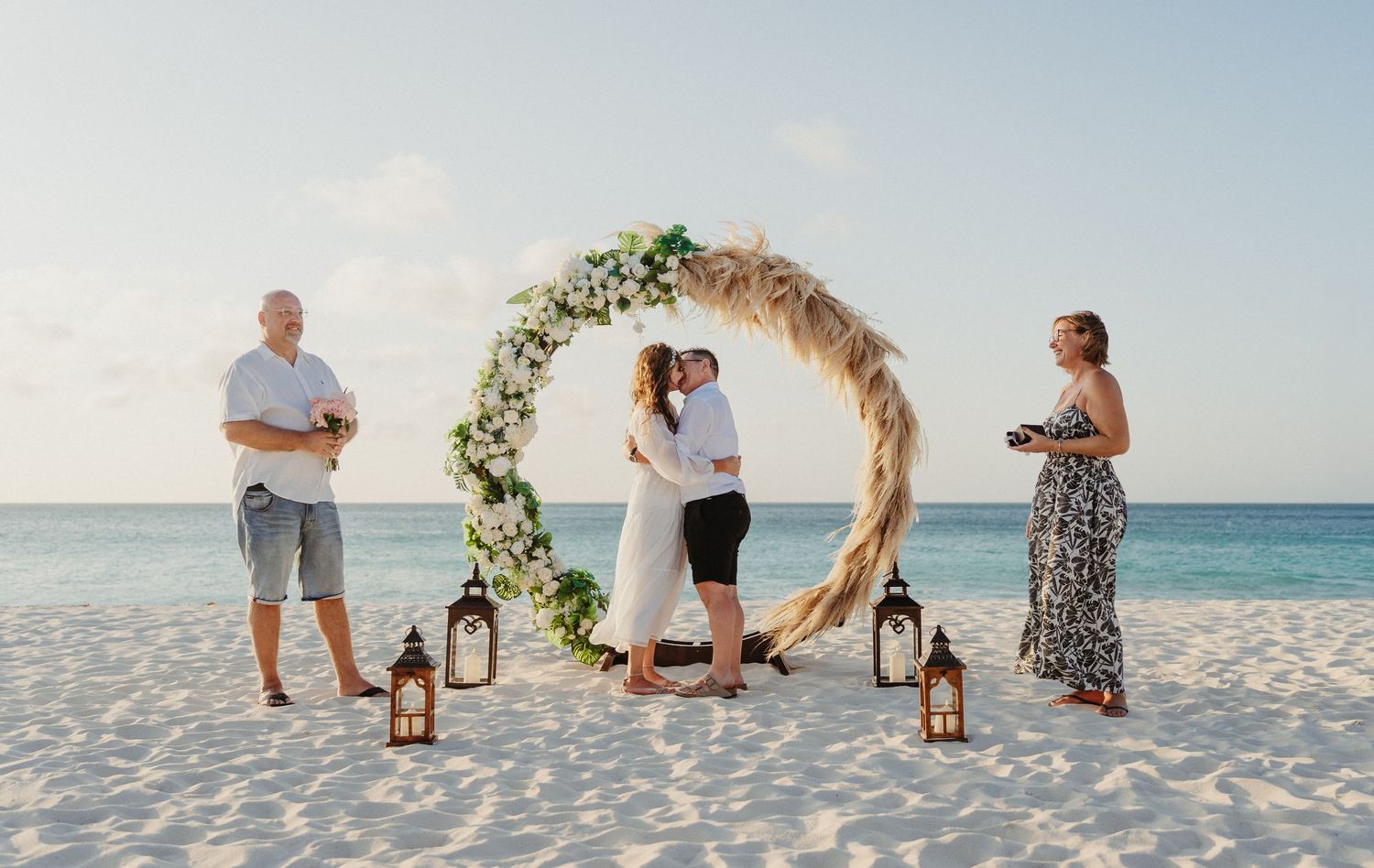 A beach wedding ceremony setup with a floral arch and lanterns on white sand overlooking turquoise waters.