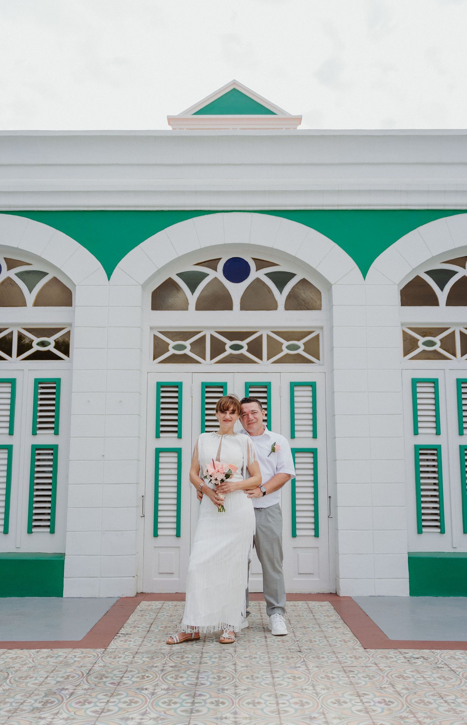 A couple stands together in front of a historic colonial building with white walls, green shutters and ornate arched windows.