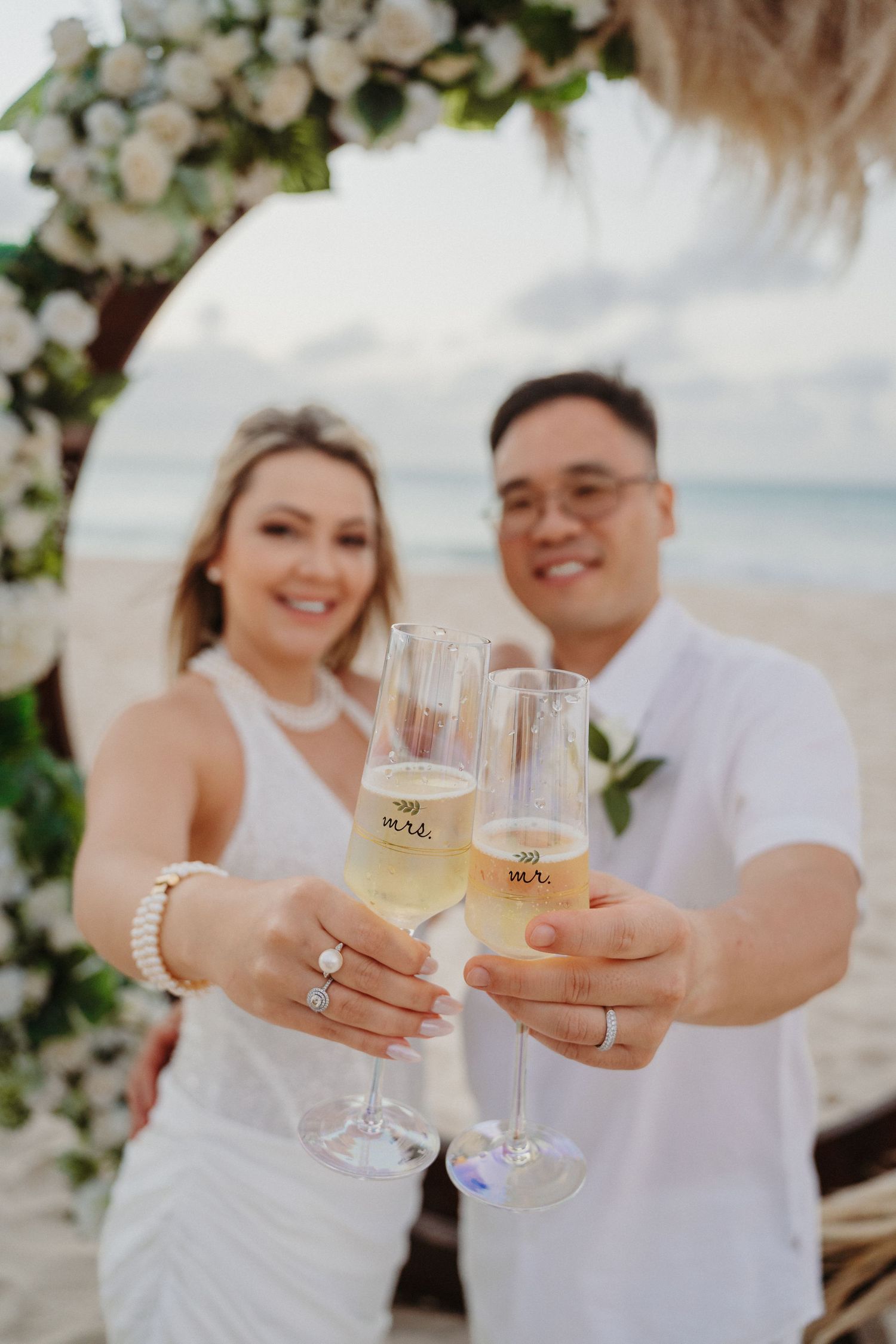 A couple toasts with champagne glasses on a beach during a romantic celebration with floral decorations.