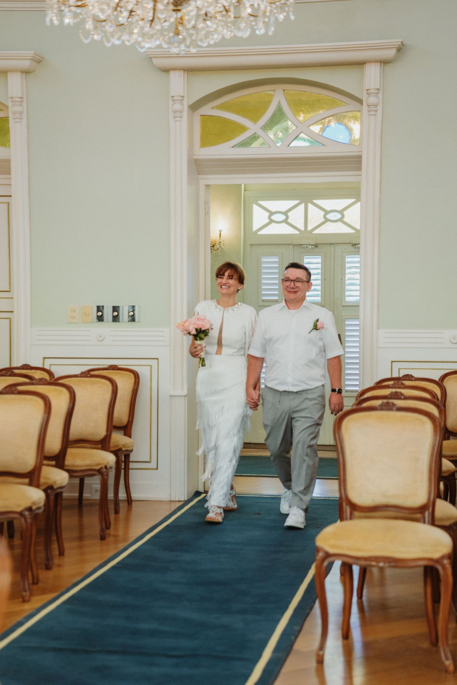A couple walks down an elegant aisle with crystal chandeliers and antique chairs in an ornate wedding ceremony venue.