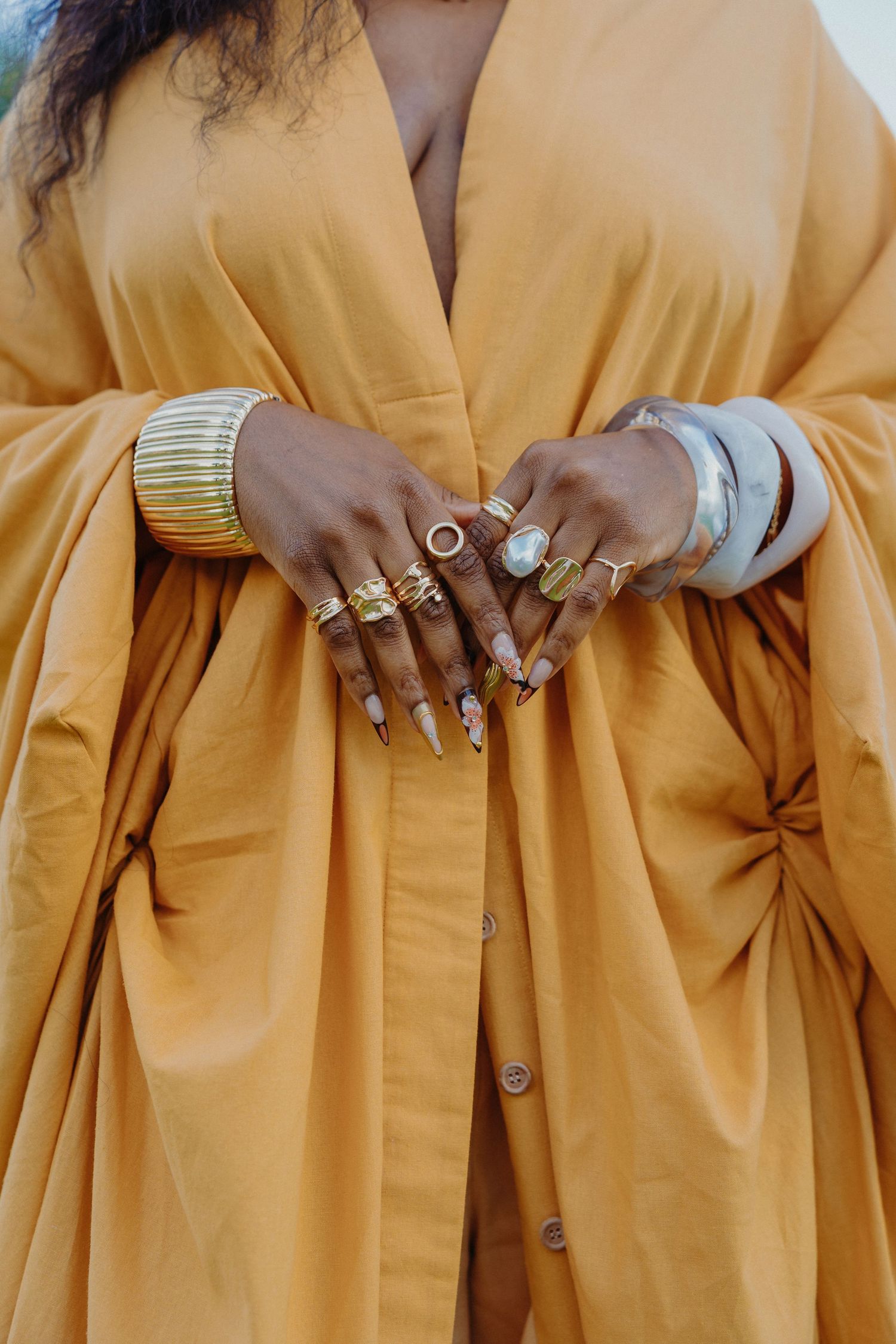 Hands adorned with multiple gold rings and bangles against a flowing mustard yellow dress.