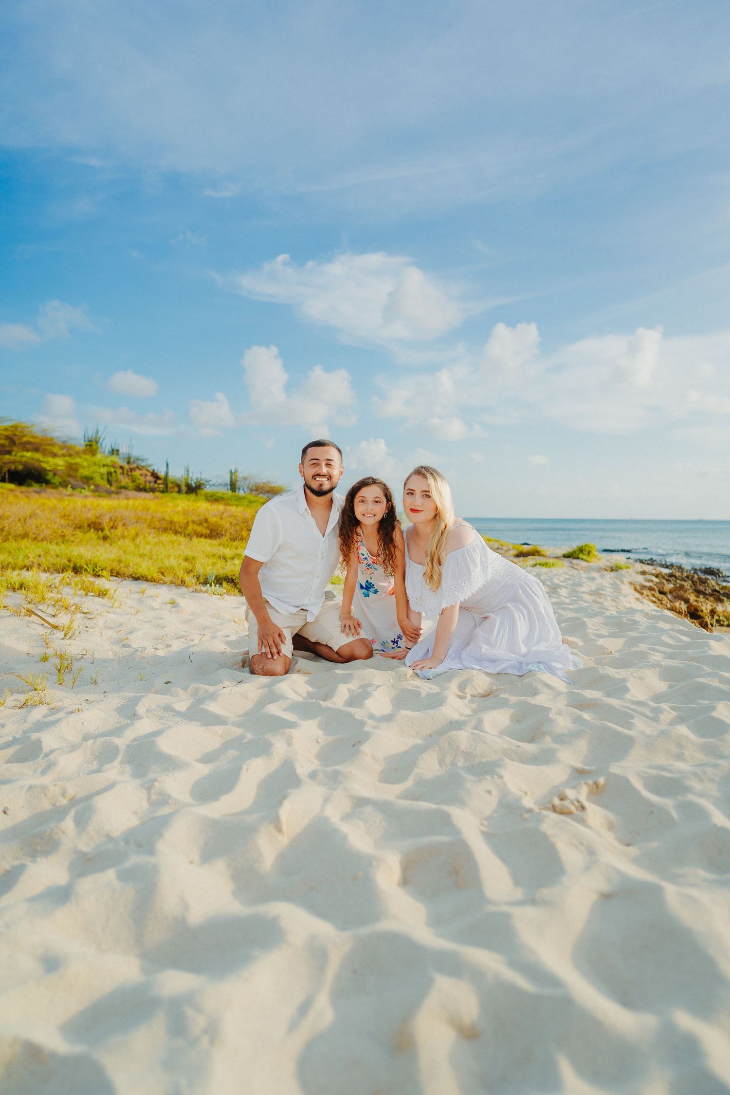 A family dressed in white poses together on a sandy beach with blue sky and ocean in the background.