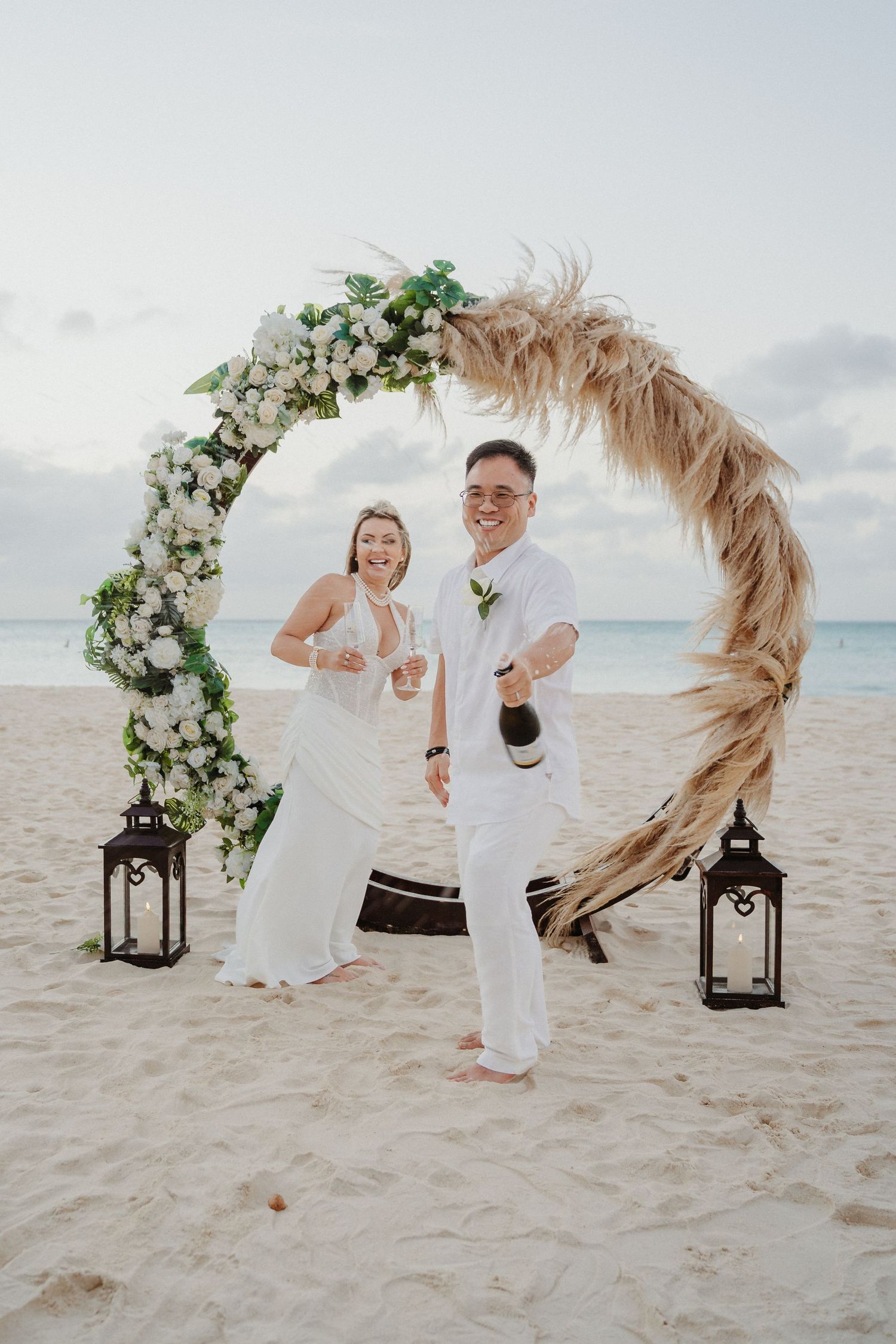A beach wedding celebration under a floral arch with lanterns on the sand by the ocean at sunset.