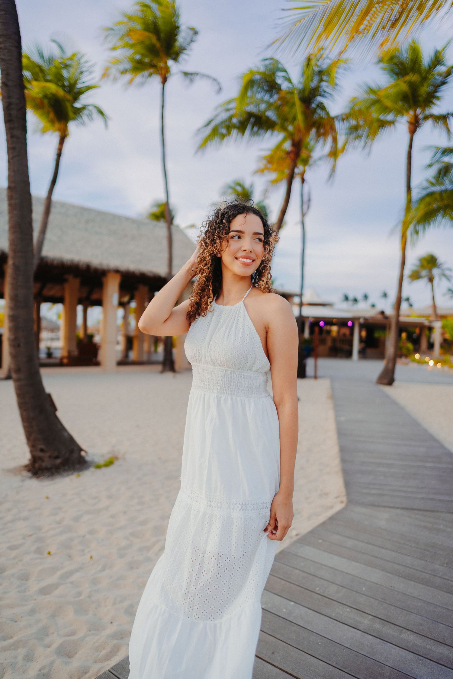 A woman in a flowing white dress walks along a beachfront boardwalk lined with palm trees on a sunny day.