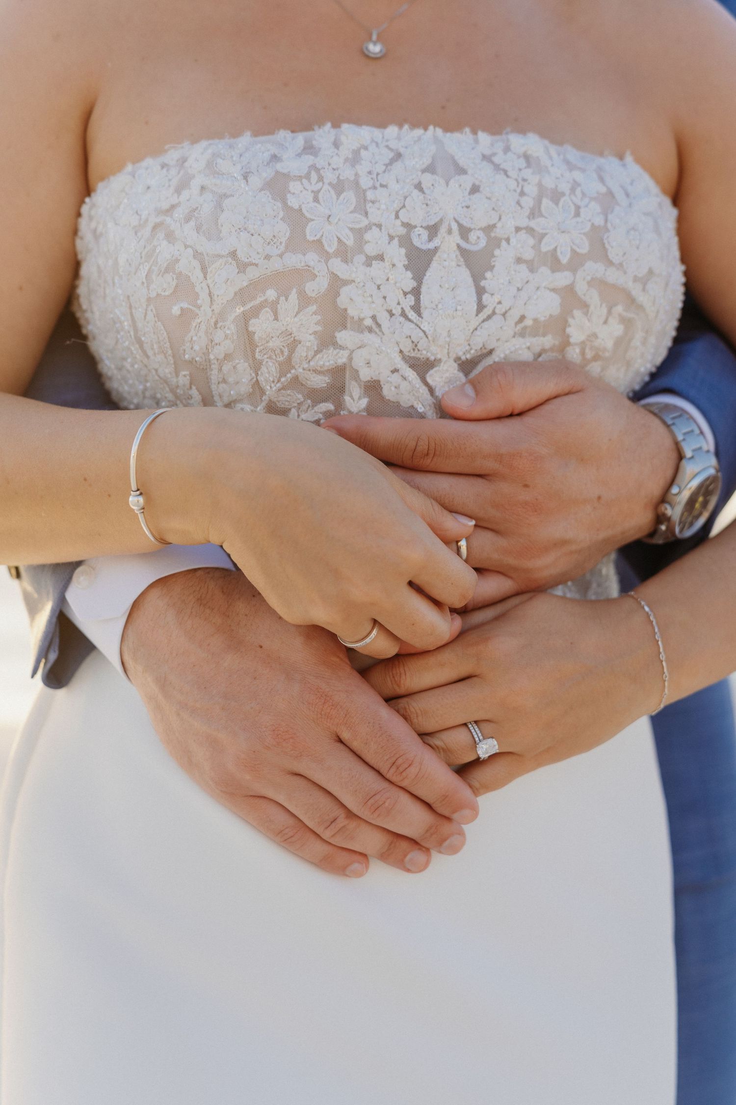 Bride and groom holding hands showing wedding rings, bride in strapless lace gown, groom in suit with watch.