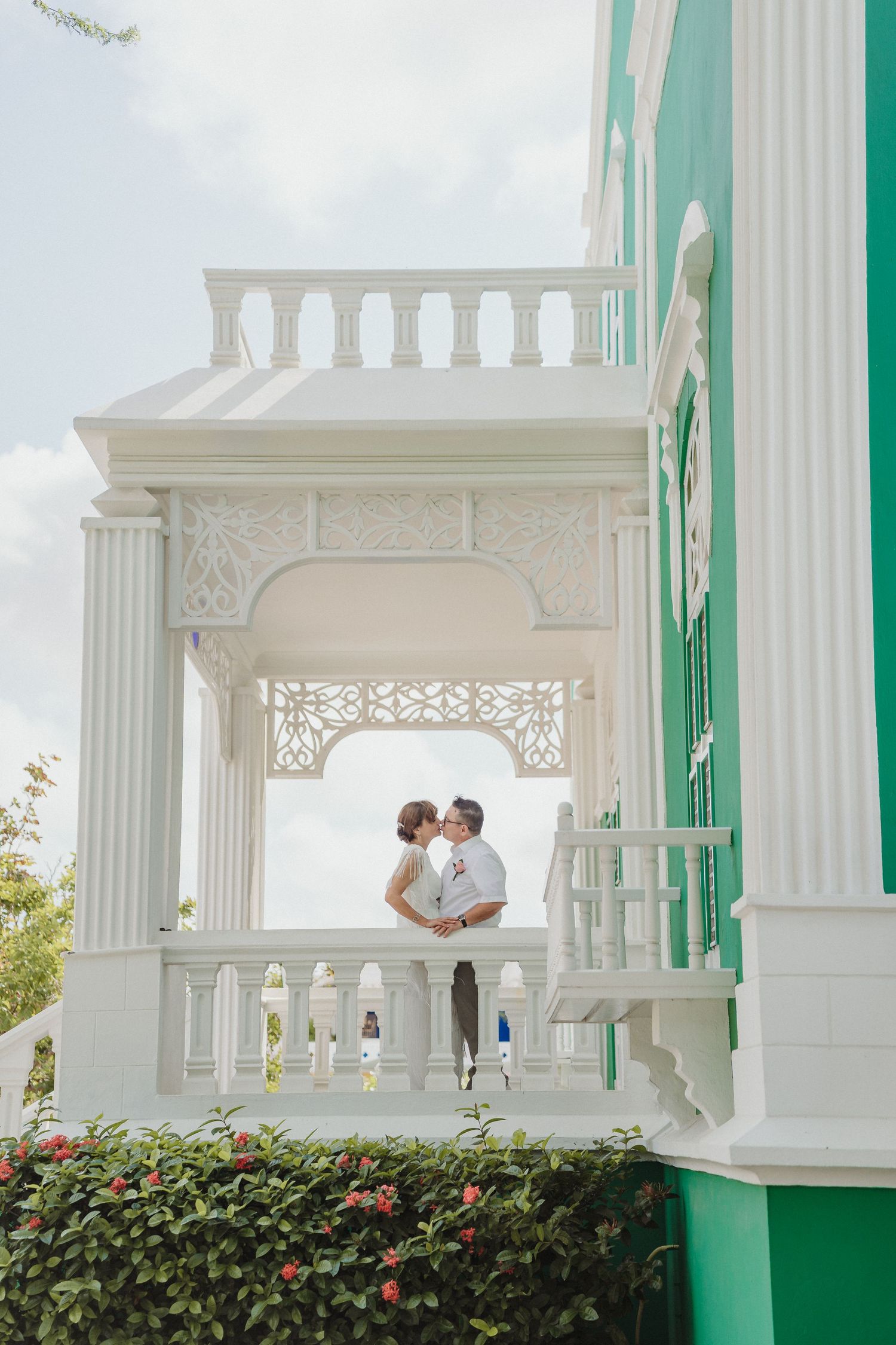 White Victorian-style porch with decorative columns and balustrades surrounded by lush greenery and flowering bushes.
