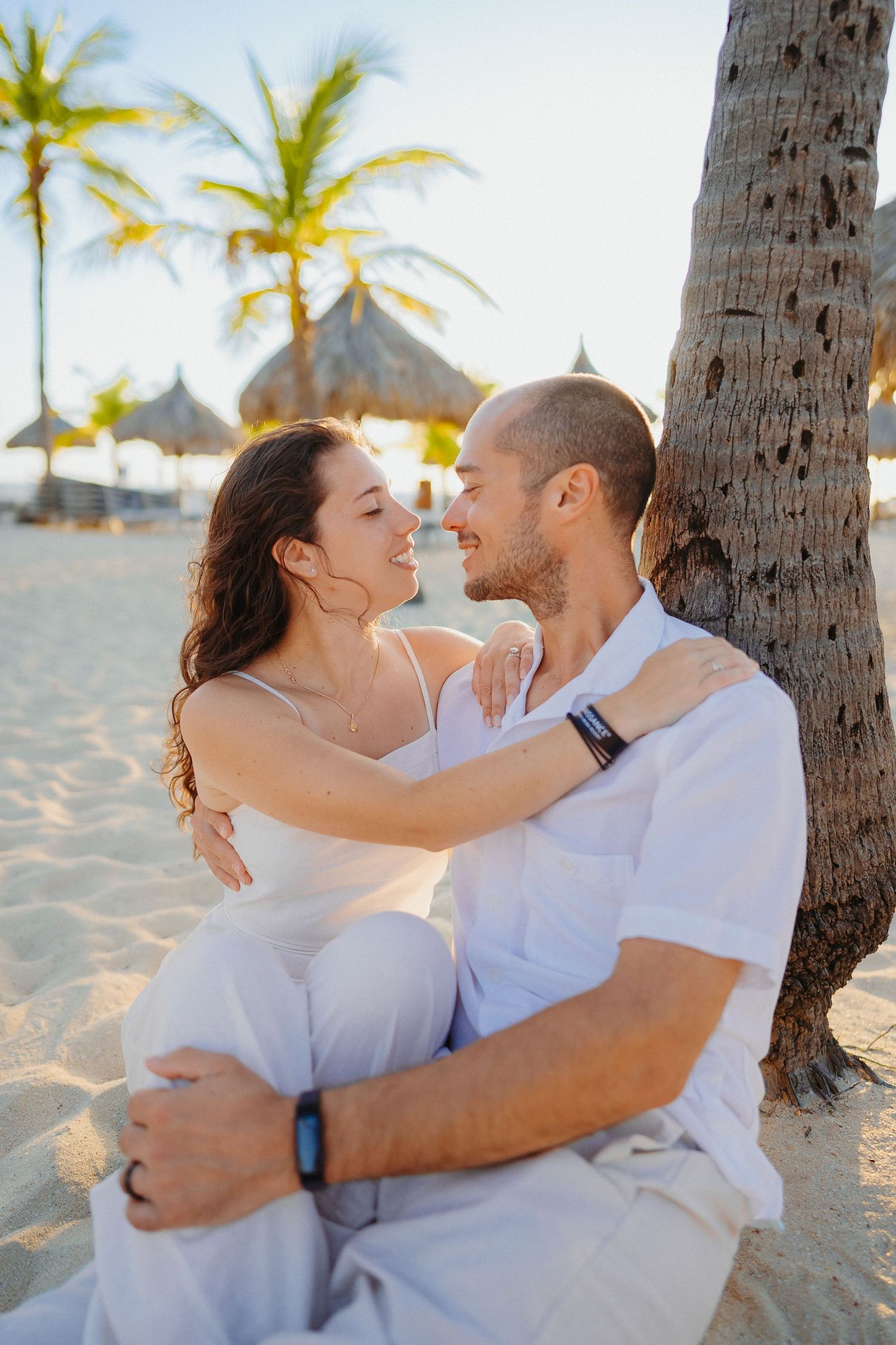 Romantic couple in white clothing embracing on a tropical beach with palm trees and thatched umbrellas at sunset.